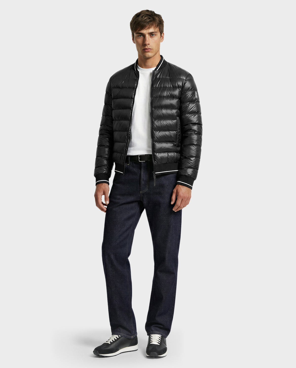 A man faces forward wearing the Rudsak JEREMY - 6125025 Black men’s down bomber jacket, a white T-shirt, dark blue jeans, and gray sneakers against a plain light gray background.