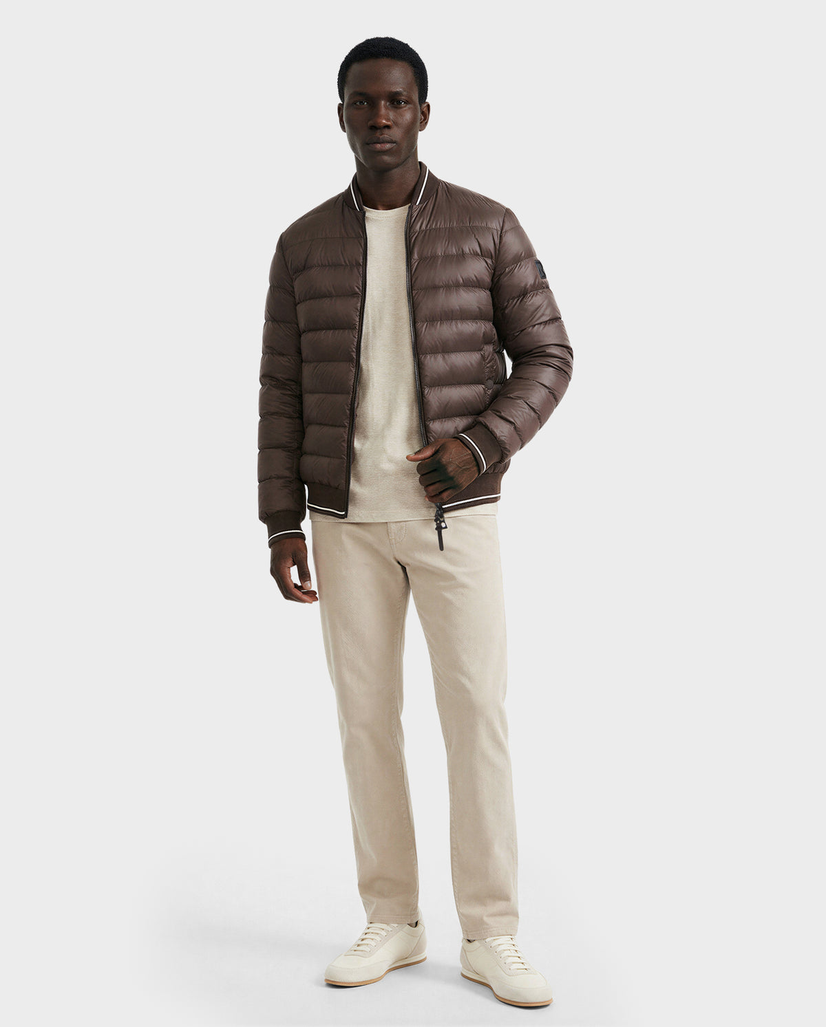 A man stands against a plain background wearing the Rudsak JEREMY - 6125025 Espresso men's down bomber jacket, paired with a light beige shirt, beige pants, and white sneakers—a cold-season staple with premium down insulation.