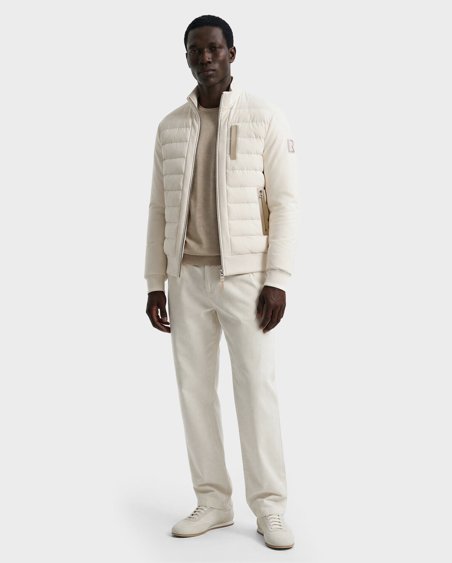 A man in the Rudsak JIMMY - 6125723 Vanilla men’s quilted down jacket, with a matching sweater, cream pants, and white sneakers stands against a plain light background.