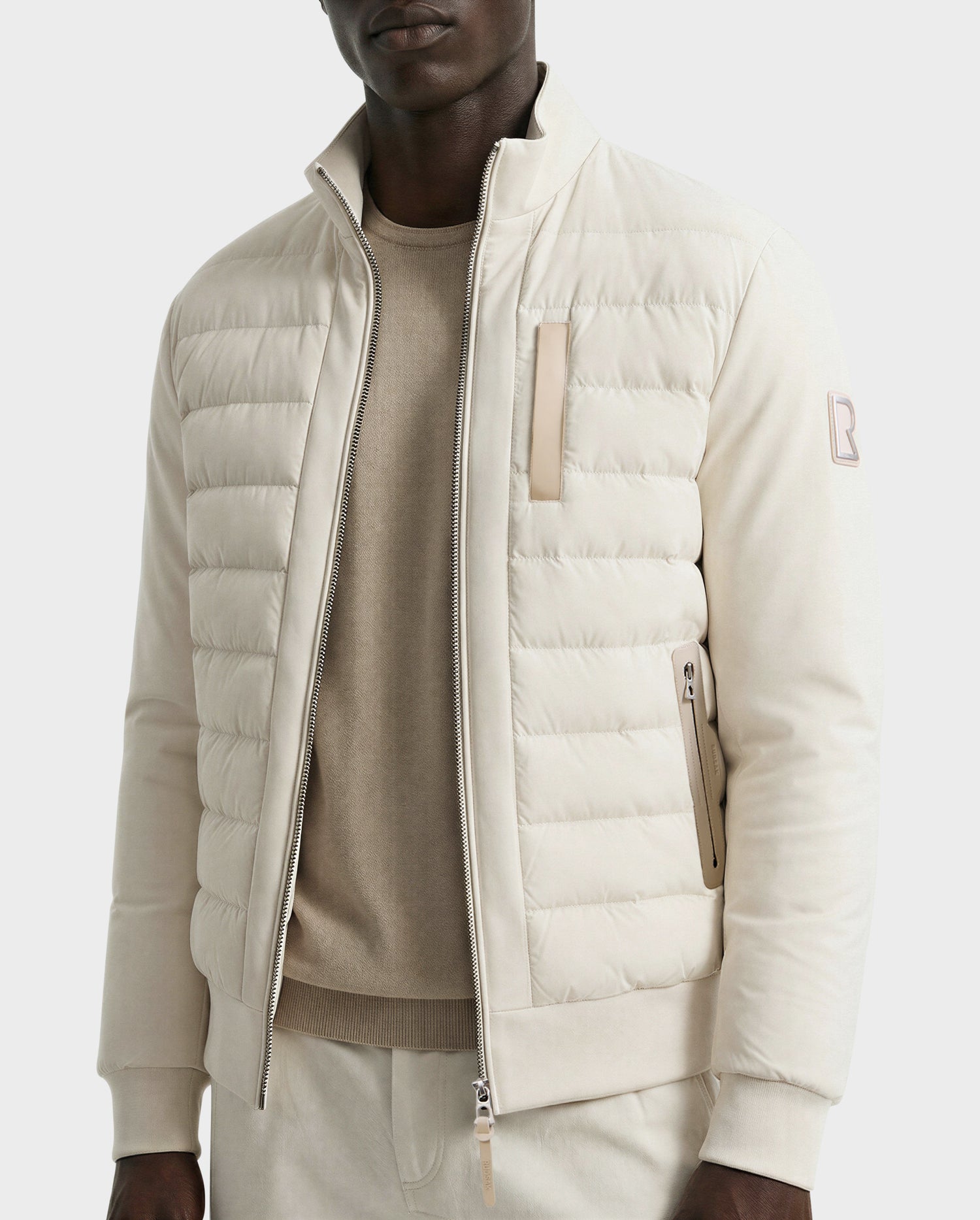 A man wears the Rudsak JIMMY - 6125723 Vanilla light beige men’s quilted down jacket with zip-up front, high collar, and zippered pockets. The water- and wind-resistant jacket is styled with a beige sweater and matching light pants.