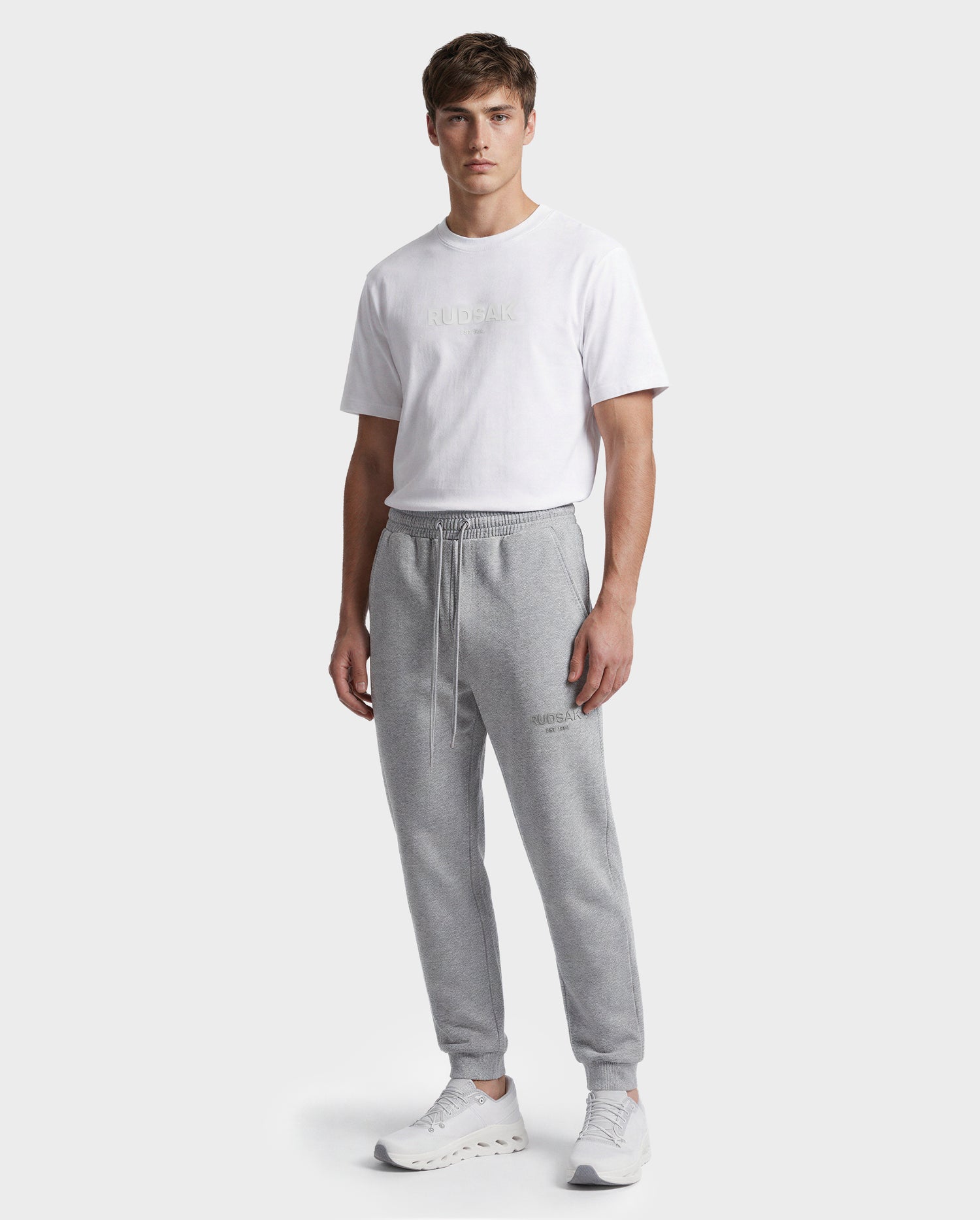 A man stands facing forward, wearing a white t-shirt, white sneakers, and Rudsak JOSS - 9524009 Heather Grey M premium cotton joggers against a plain light gray background.