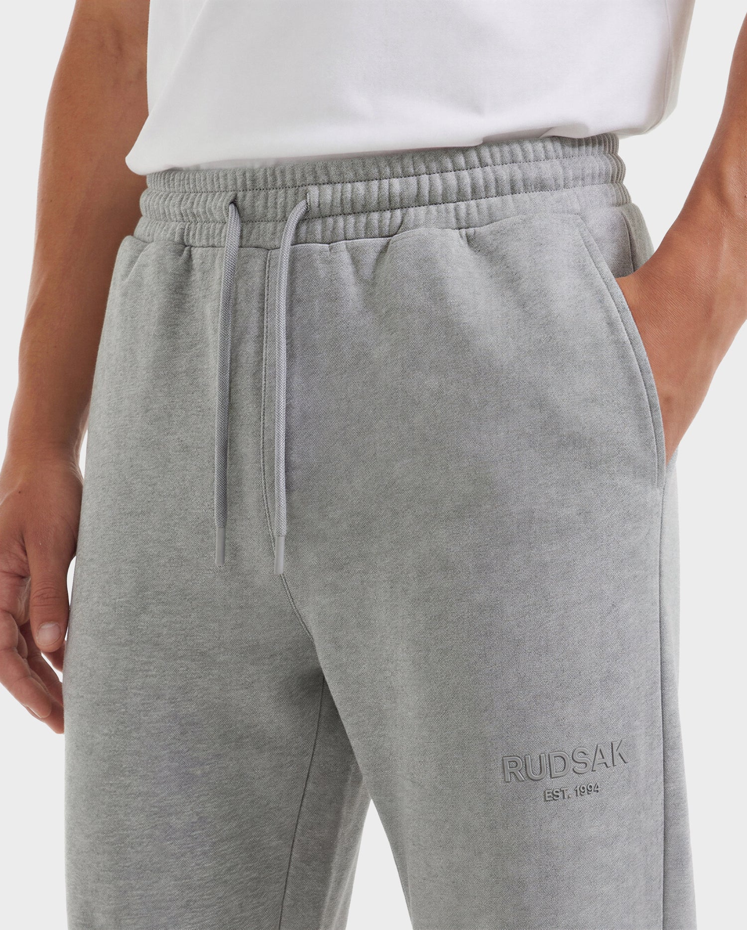 A person wearing Rudsak JOSS - 9524009 joggers in heather grey (M), made from premium cotton with an elastic waistband, drawstring, side pocket, and RUDSAK EST. 1994 embroidery on the left thigh, paired with a white t-shirt.
