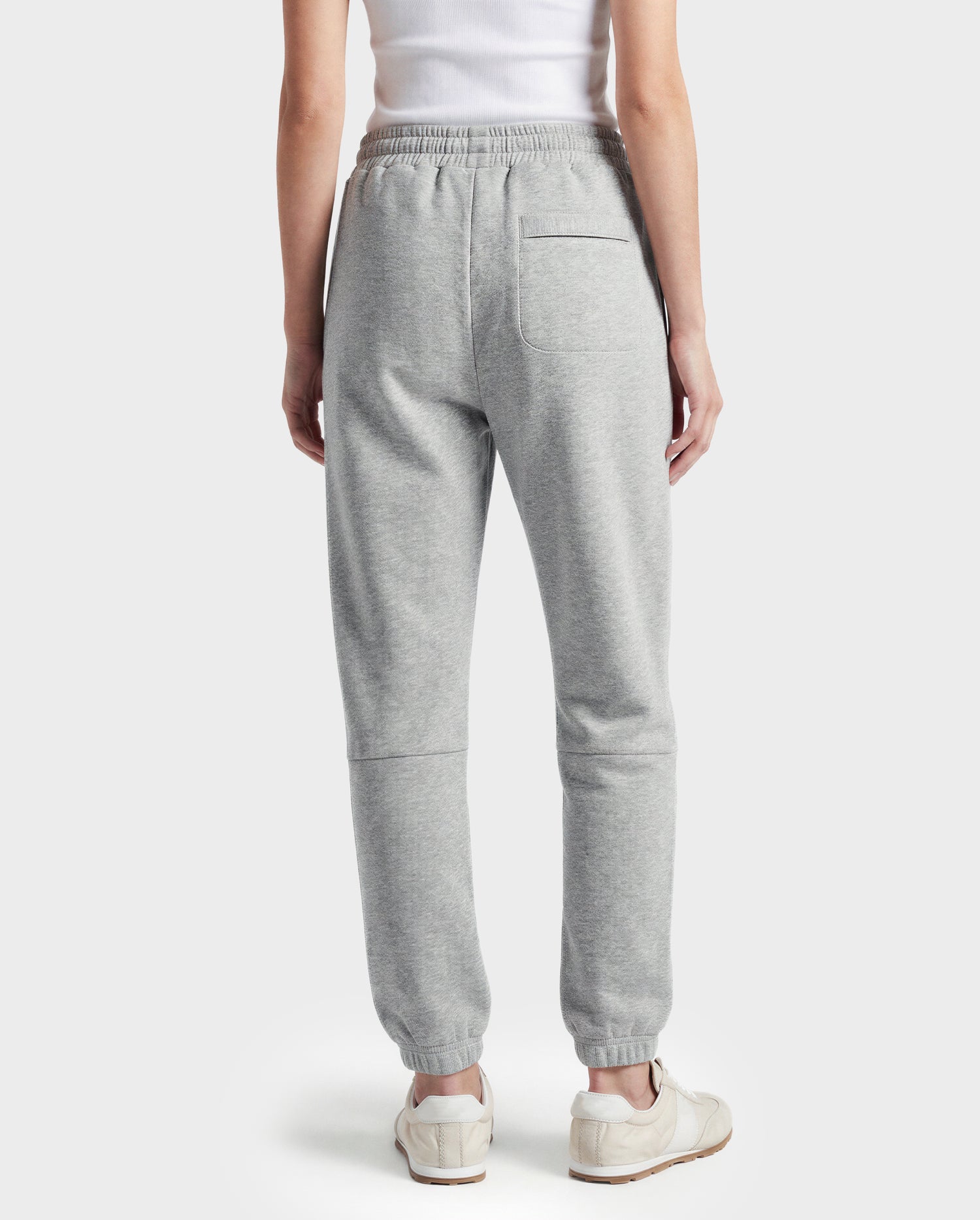 A person stands facing away, wearing white sneakers, a white tank top, and Rudsak's JOSS - 9524009 Heather Grey W premium joggers, showcasing the back of this stylish look against a plain light background.