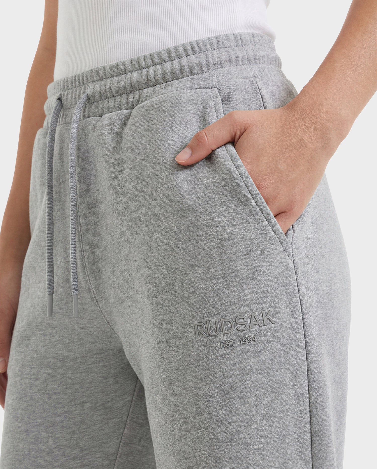 A person wearing Rudsak's JOSS - 9524009 Heather Grey W joggers with RUDSAK EST. 1994 embroidered on the pocket and a white ribbed tank top stands with one hand in their pocket.