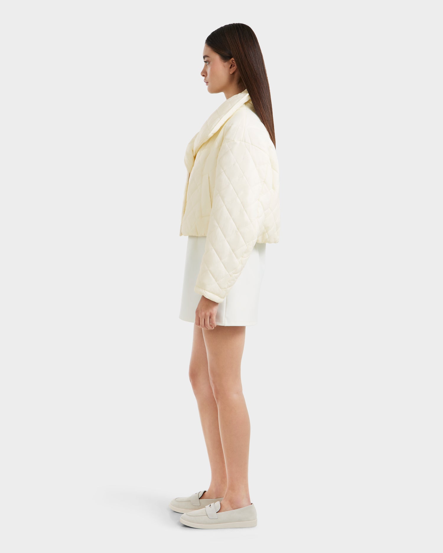 A woman stands in profile wearing the Rudsak KALEE - 8124005 Iceland quilted light jacket, a white mini skirt, and light-colored loafers against a plain light gray background.