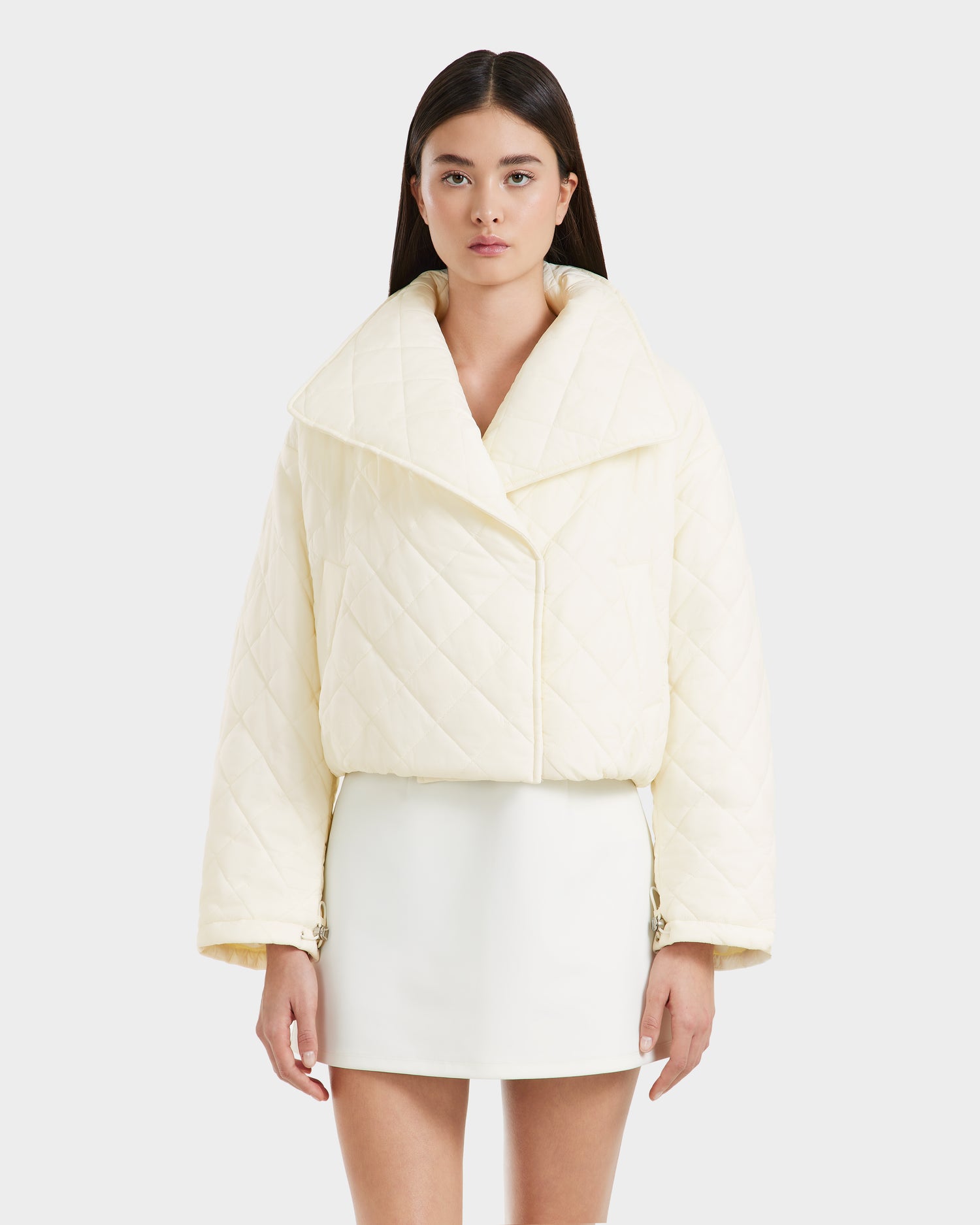 A woman with straight dark hair wears the Rudsak KALEE - 8124005 Iceland quilted jacket with a large collar, paired with a white mini skirt, standing against a plain light background.
