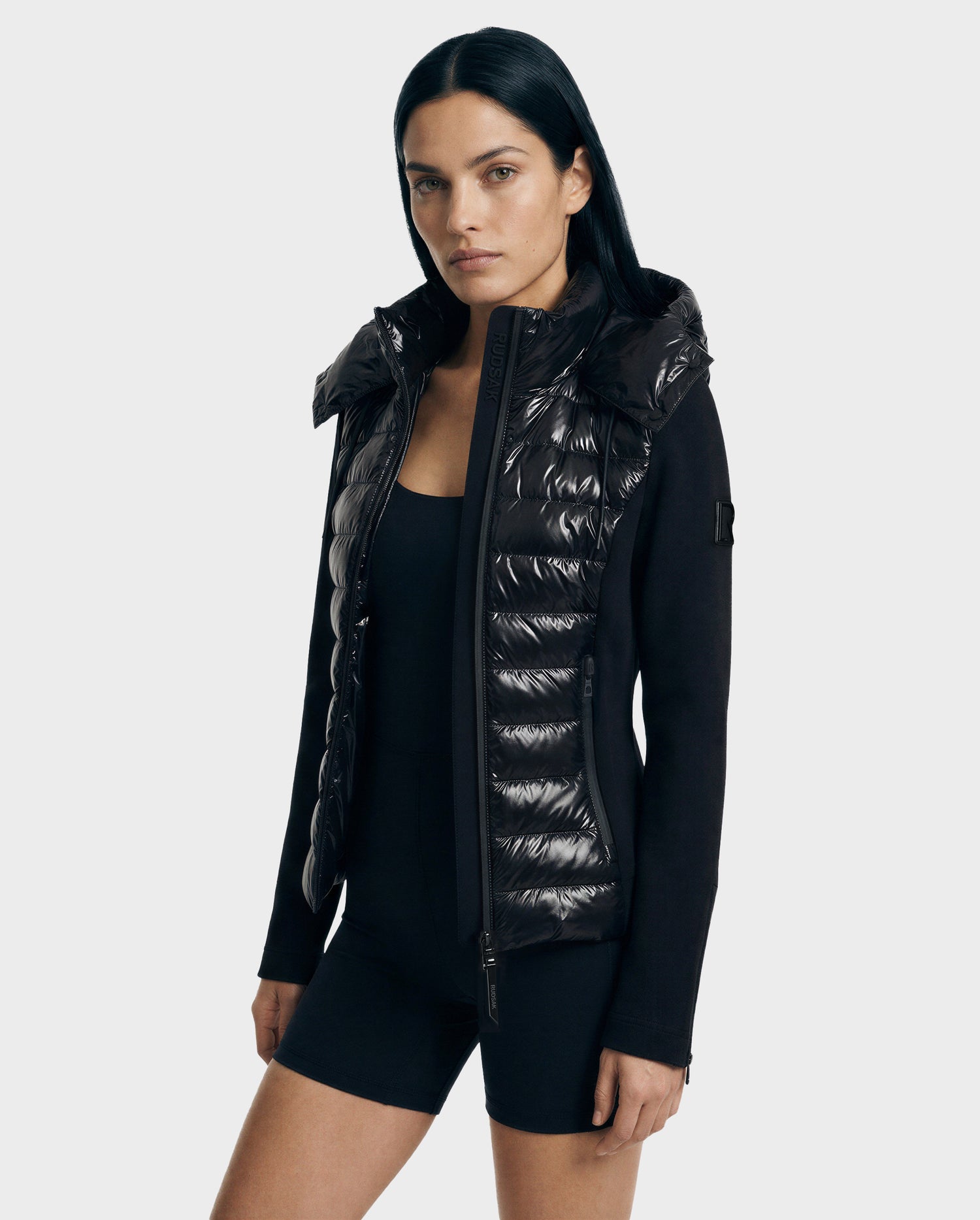 A woman with straight black hair wears the Rudsak MABS C - 8125701 Black puffer jacket and black athletic shorts, standing against a plain light background.