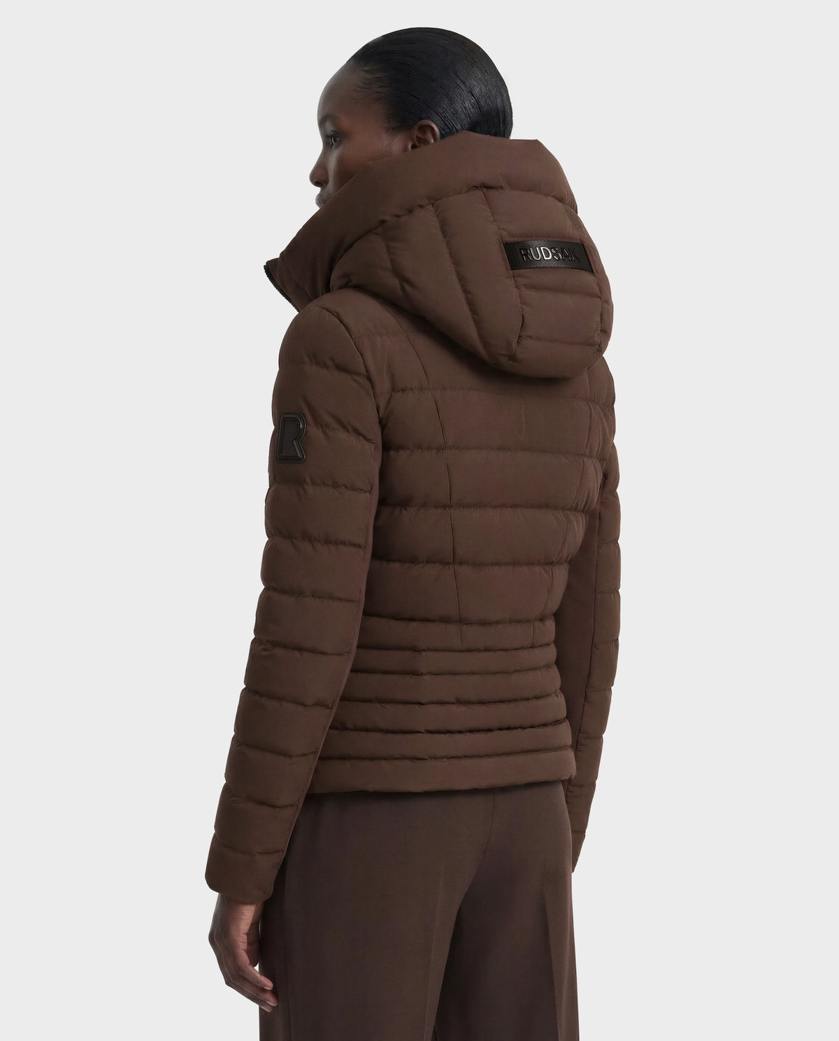 A person faces away, displaying the back and side of the Rudsak MELISSA - 8124824 Espresso women's lightweight down puffer with a large hood and patch details, set against a plain white background.