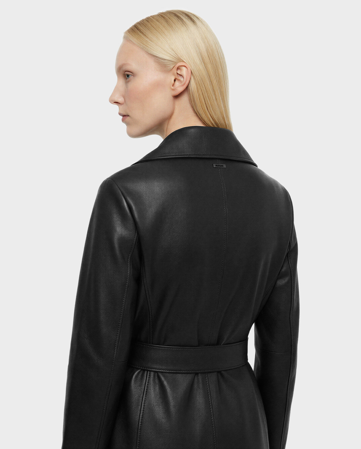 A person with straight, blonde hair models the Rudsak MURMU X - 8125670 Black leather coat with a belt from the back and side against a plain light gray background—an ideal investment piece.