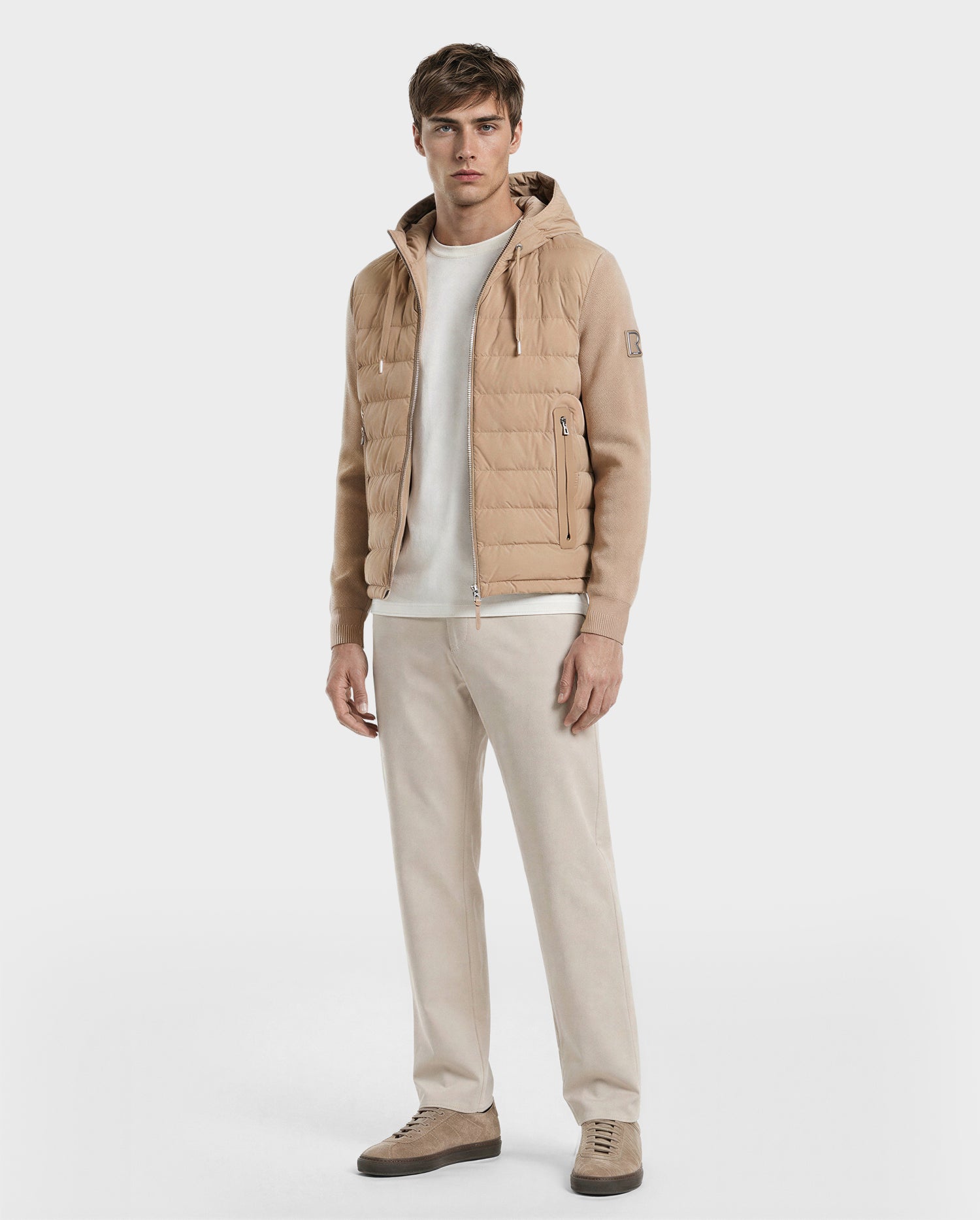 A man with short brown hair and a neutral expression stands against a plain white background wearing the RIVER - 6124584 Almond men's lightweight down jacket by Rudsak over a white shirt, beige pants, and beige sneakers.