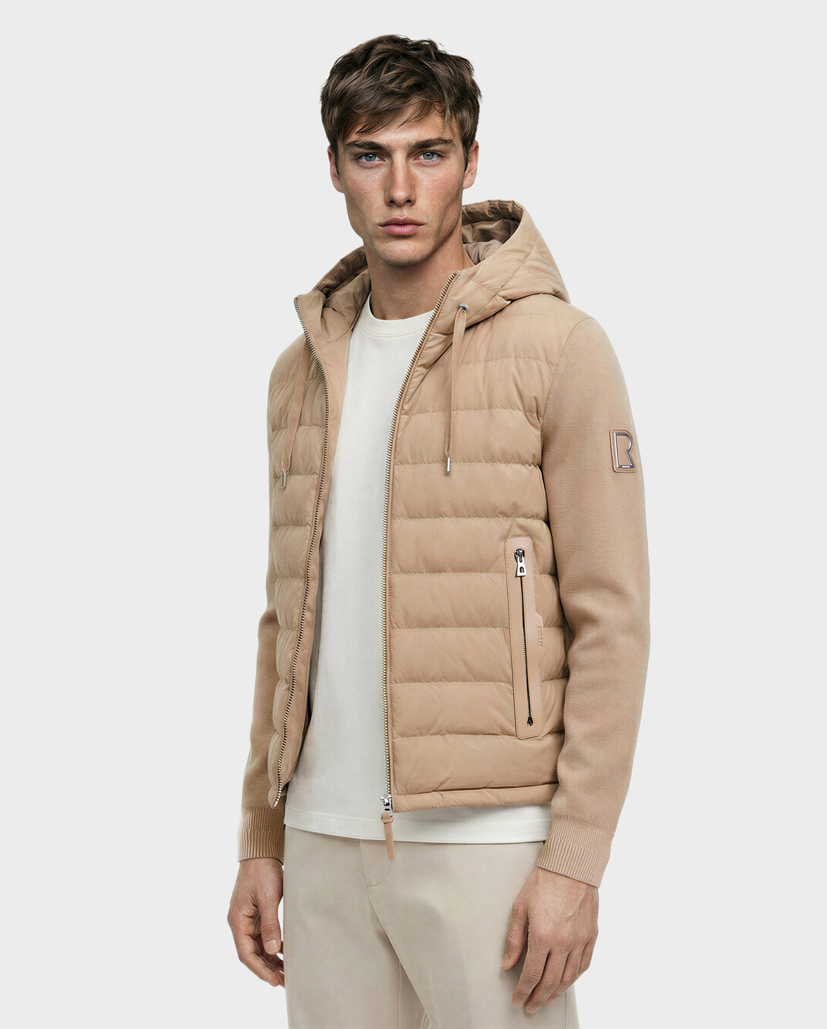 A young man stands facing forward against a plain, light background, wearing the Rudsak RIVER - 6124584 Almond beige hooded down jacket over a white T-shirt and beige pants.