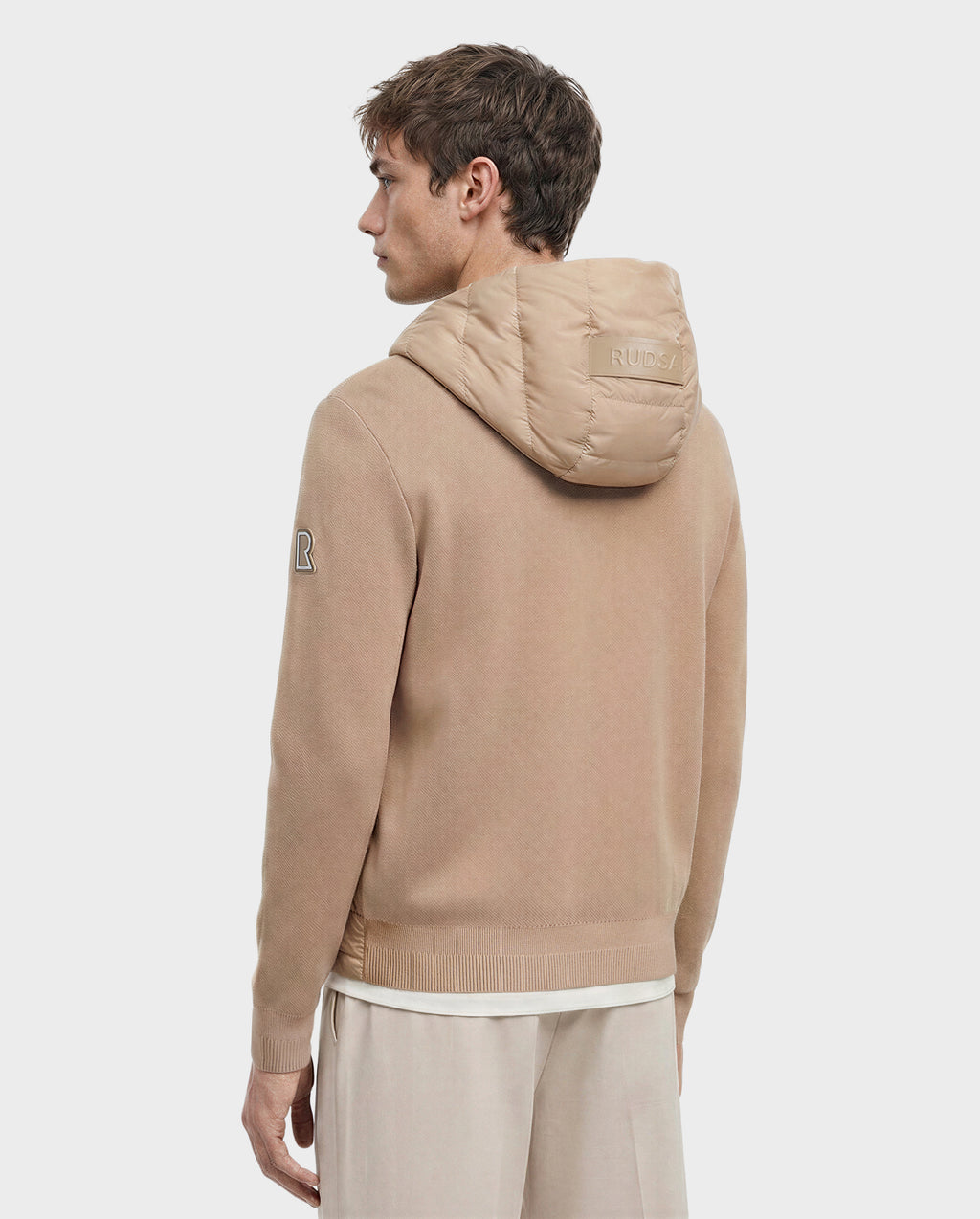 A man with light brown hair faces away, wearing the Rudsak RIVER - 6124584 Almond hooded down jacket with "RUDSAK" on the hood and an "R" patch on the sleeve, paired with light pants against a plain white background.