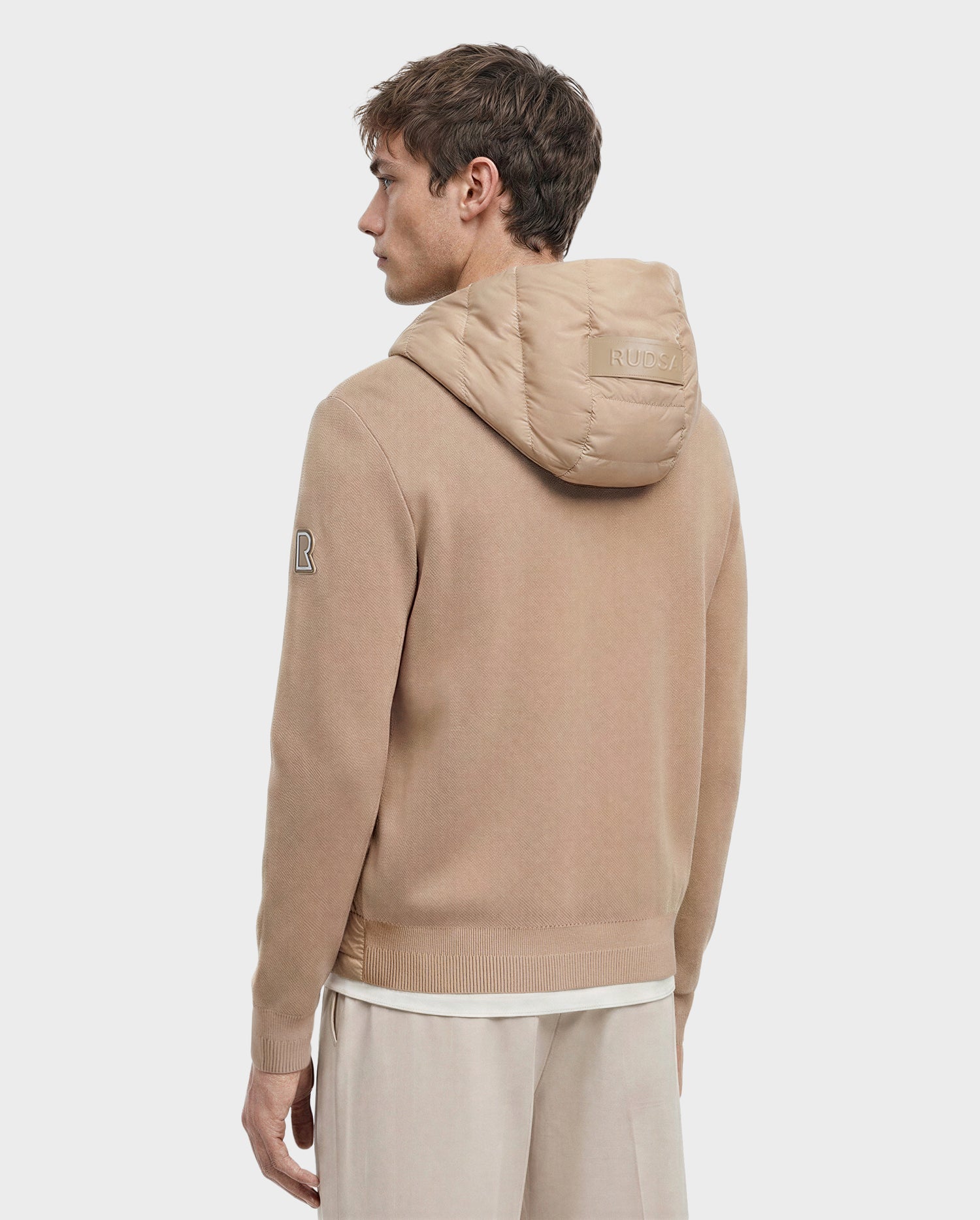 A man with light brown hair faces away, wearing the Rudsak RIVER - 6124584 Almond hooded down jacket with "RUDSAK" on the hood and an "R" patch on the sleeve, paired with light pants against a plain white background.