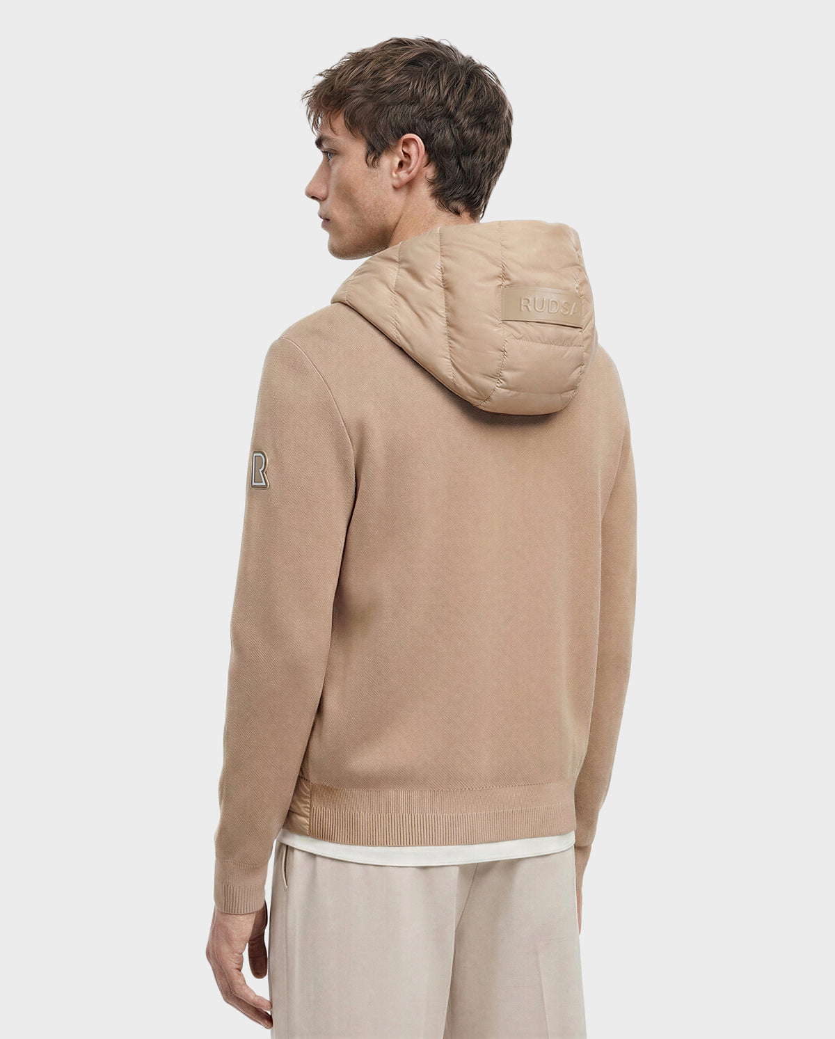 A man with light brown hair faces away, wearing the Rudsak RIVER - 6124584 Almond hooded down jacket with "RUDSAK" on the hood and an "R" patch on the sleeve, paired with light pants against a plain white background.