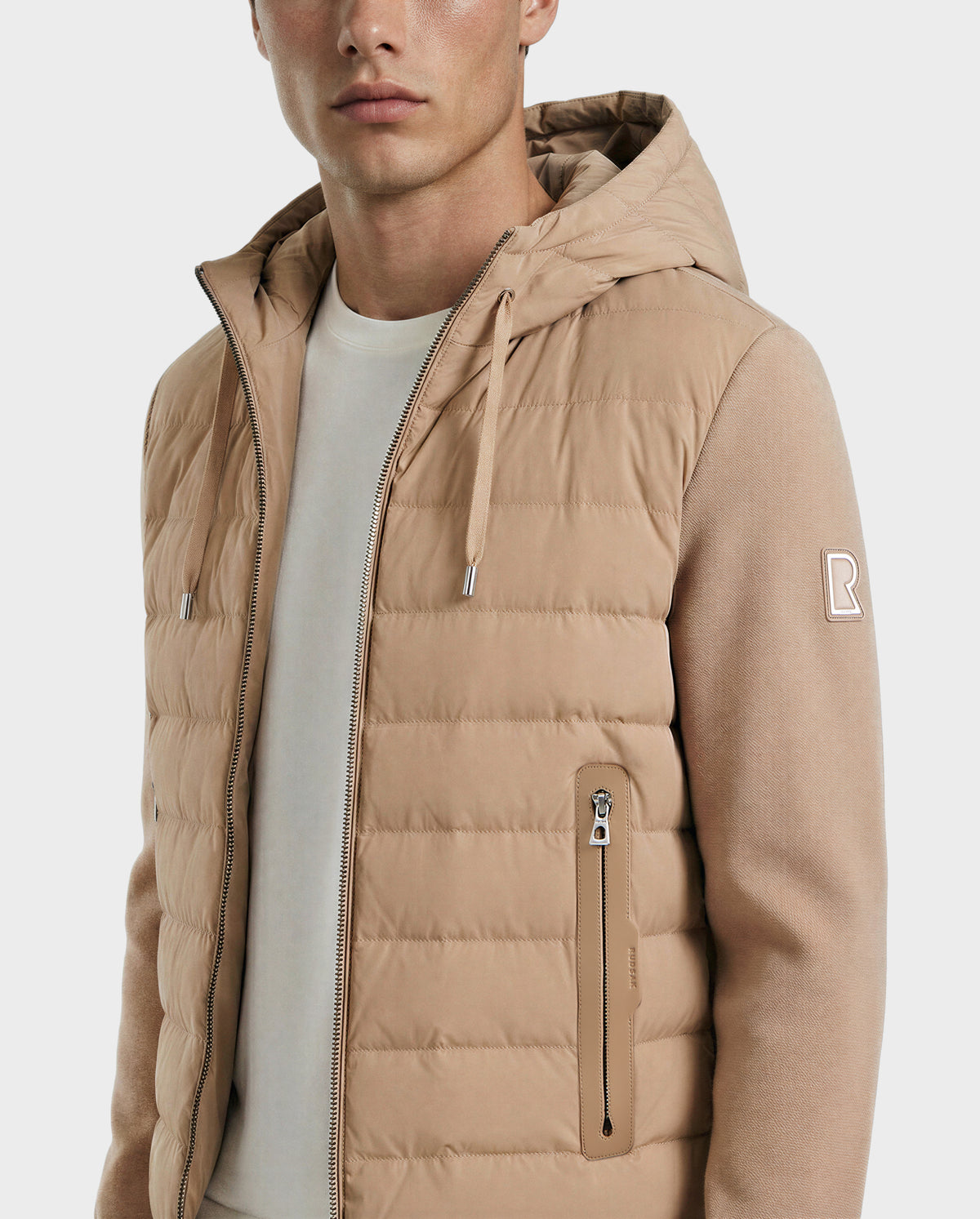 A man wears the Rudsak RIVER - 6124584 Almond men's lightweight down jacket with ribbed sleeves and zippered pockets over a white t-shirt, shown from the chest up against a plain background.