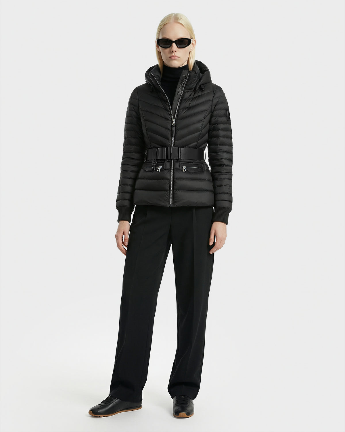 A person wearing Rudsak’s SCARLETT - 8125004 Black women’s lightweight down jacket with a vegan leather waist belt, black turtleneck, black pants, black shoes, and sunglasses stands against a plain white background.