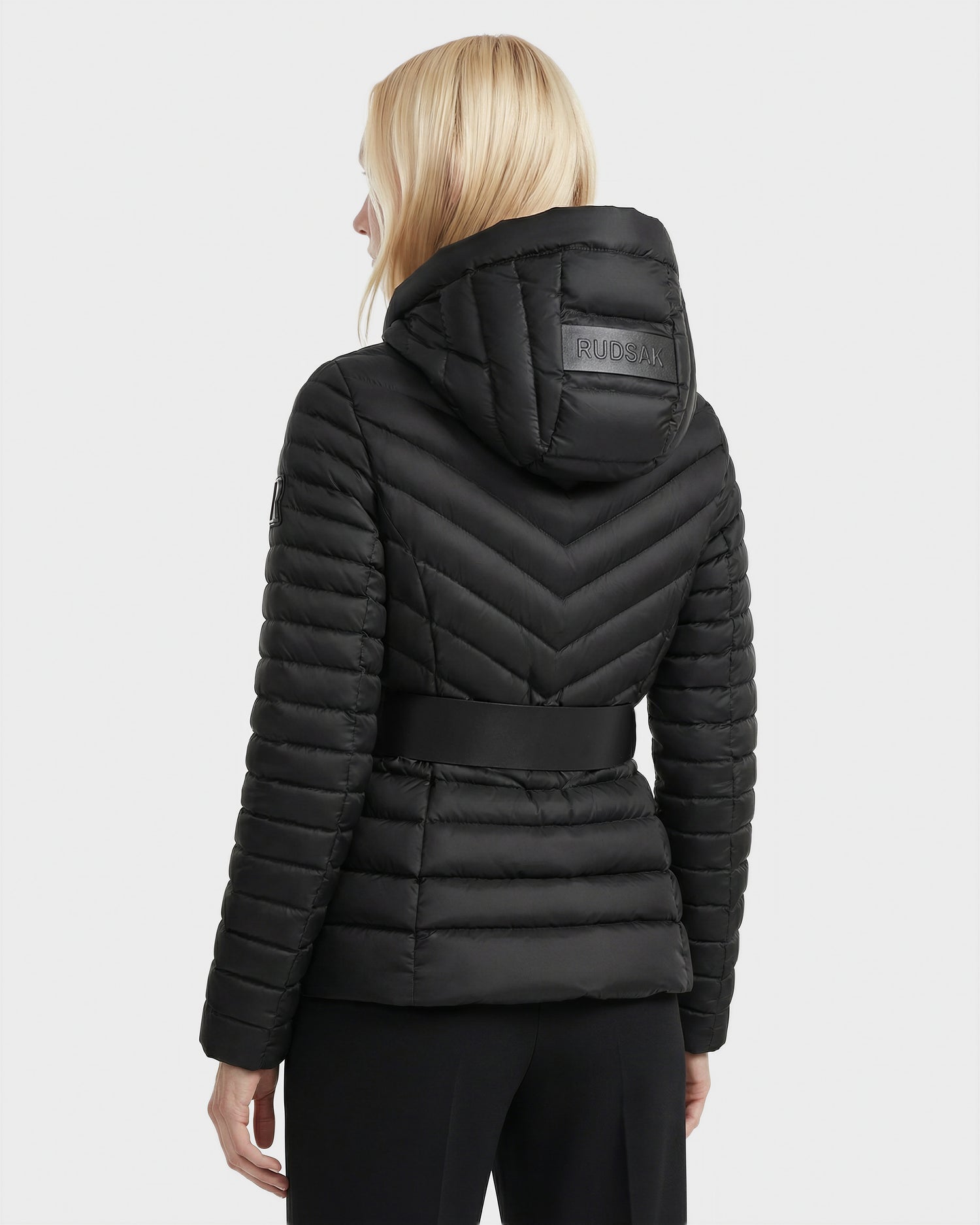 A woman with blonde hair, facing away, wears the Rudsak SCARLETT - 8125004 Black puffer jacket featuring water-resistant fabric, horizontal and chevron quilting, a wide waist belt, vegan leather trim, and branded hood detailing.