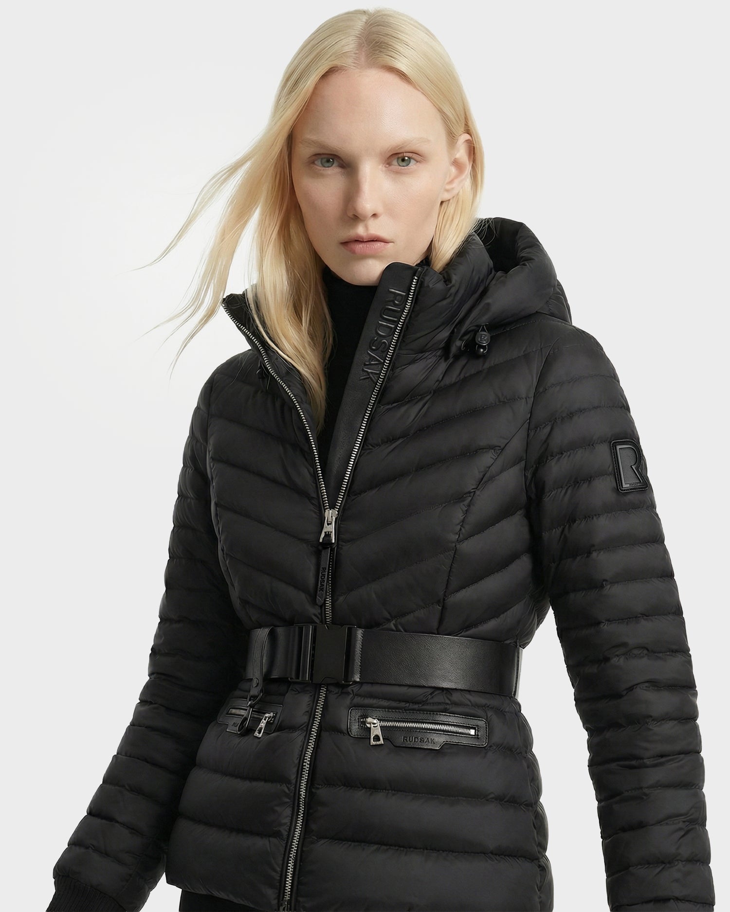 A person with long blonde hair wears the Rudsak SCARLETT - 8125004 Black water-resistant quilted puffer jacket with a high collar, zip front, vegan leather waist belt, and zippered pockets against a plain light background.