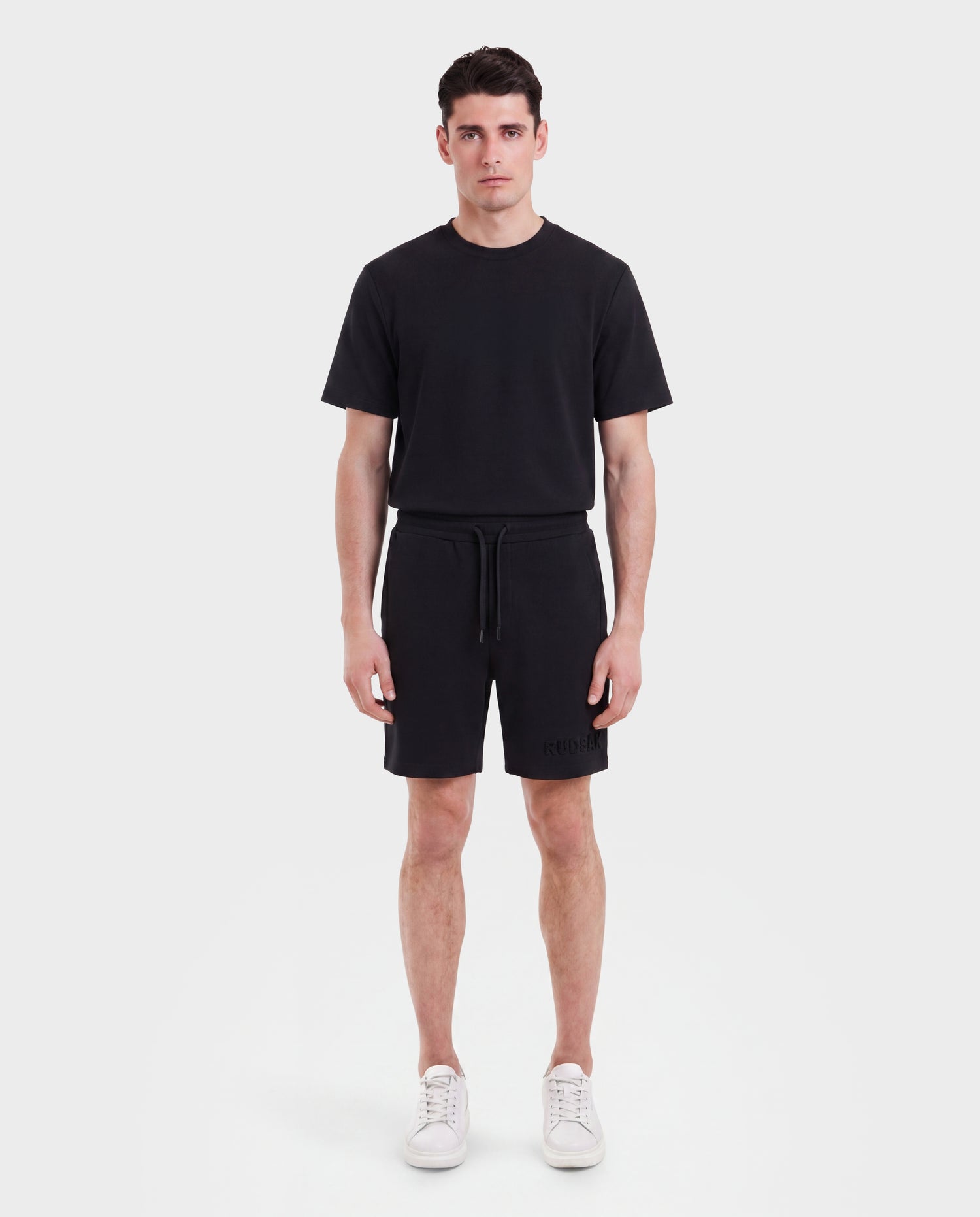 A man stands facing forward against a plain white background, wearing the Rudsak TRAVIS - M525002 Black t-shirt with sporty-casual shorts and white sneakers. His arms are relaxed by his sides—perfect for everyday wear.