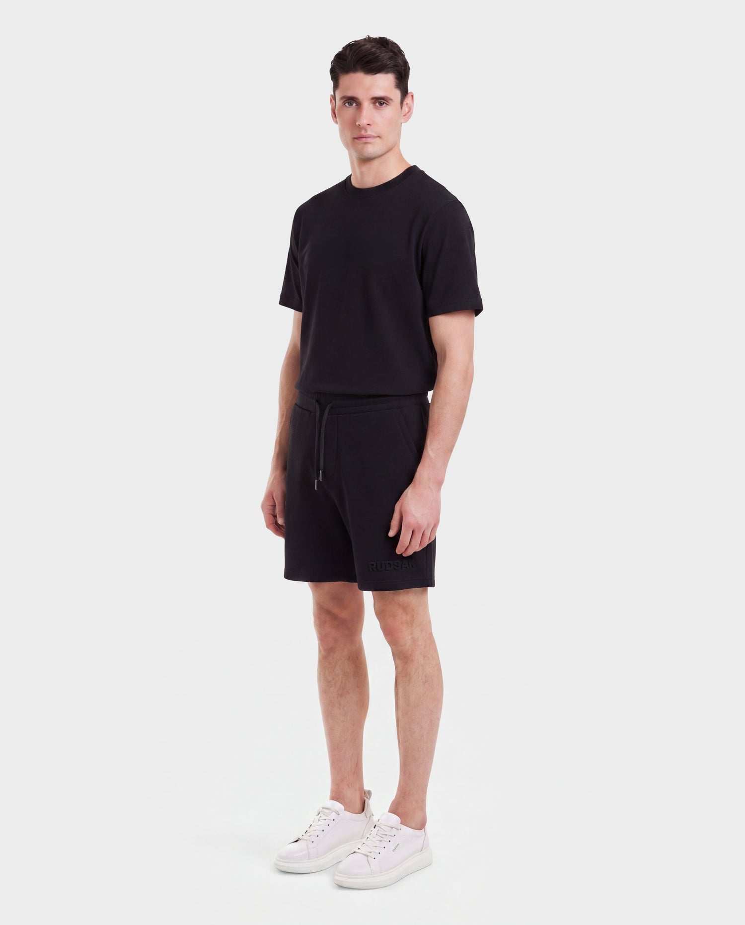 A man faces forward in a Rudsak TRAVIS - M525002 Black t-shirt, sporty-casual drawstring shorts, and white sneakers, set against a white background.