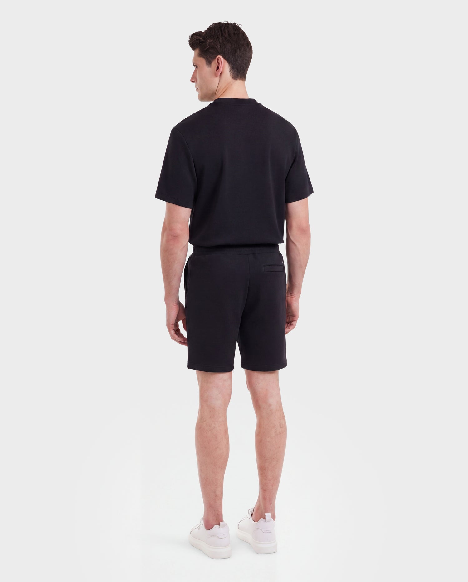 A man stands with his back to the camera, wearing the Rudsak TRAVIS - M525002 Black t-shirt, sporty-casual shorts, and white sneakers against a plain white background.