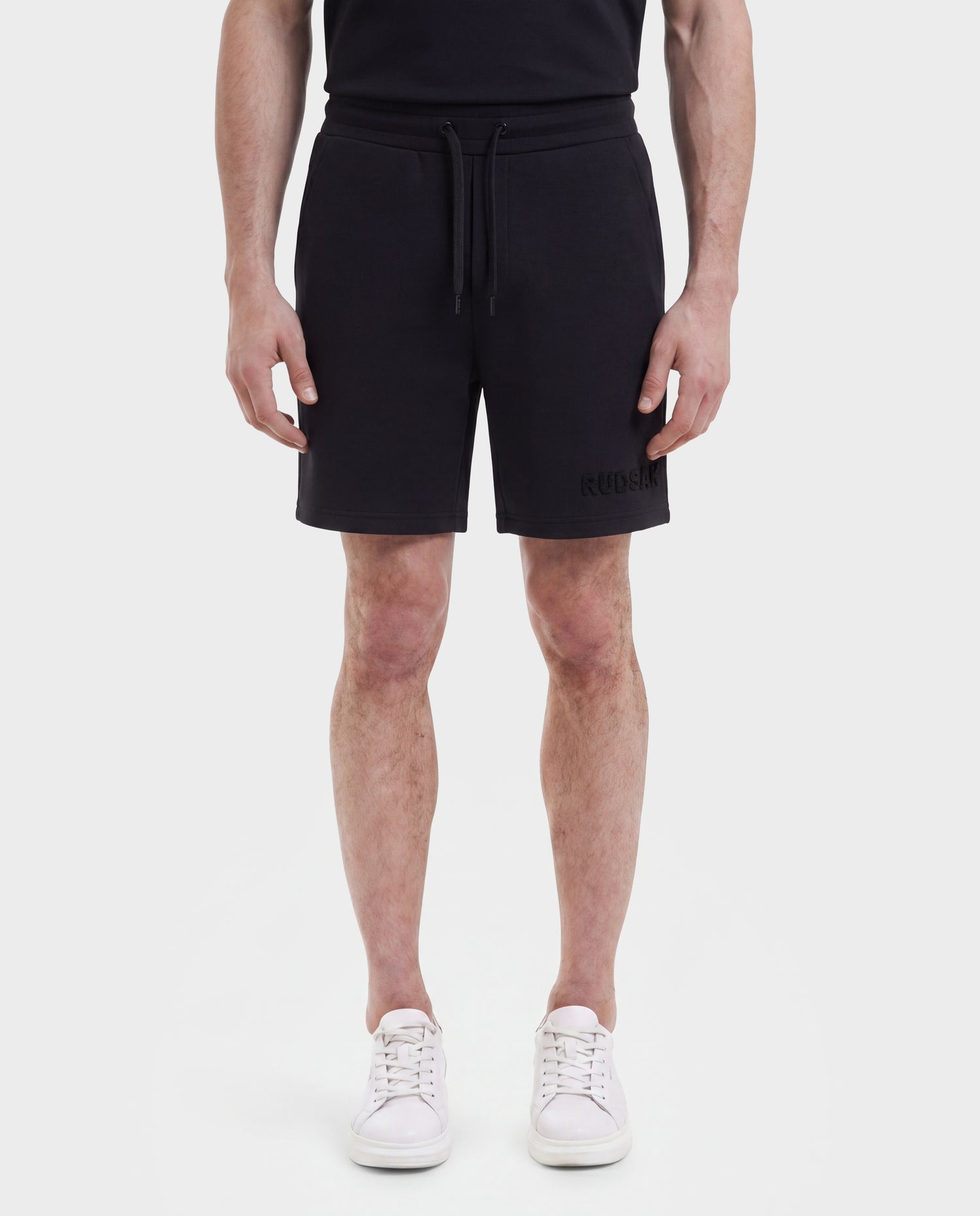 A person is shown from shoulders to feet, facing forward against a plain white background, wearing Rudsak TRAVIS - M525002 Black t-shirt, sporty-casual shorts, and white sneakers.
