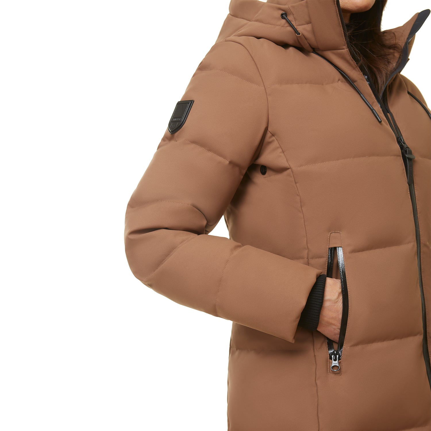 A person wears the Rudsak SYDNEY COM - 4123511 Taupe puffer jacket, featuring water-repellent fabric, black zippers, a hood, a patch on the upper sleeve, and one hand in a zippered pocket. The person's head is partially out of frame.
