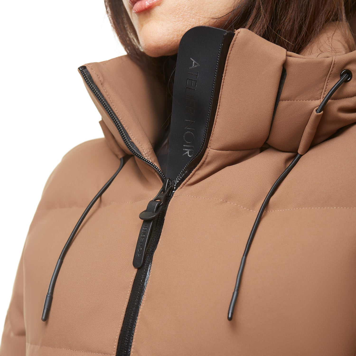 Close-up of a person wearing the Rudsak SYDNEY COM - 4123511 Taupe wind-resistant puffer with black drawstrings and inner collar; the partially unzipped duck-down parka reveals "ATELIER NOIR" text, face mostly out of frame.
