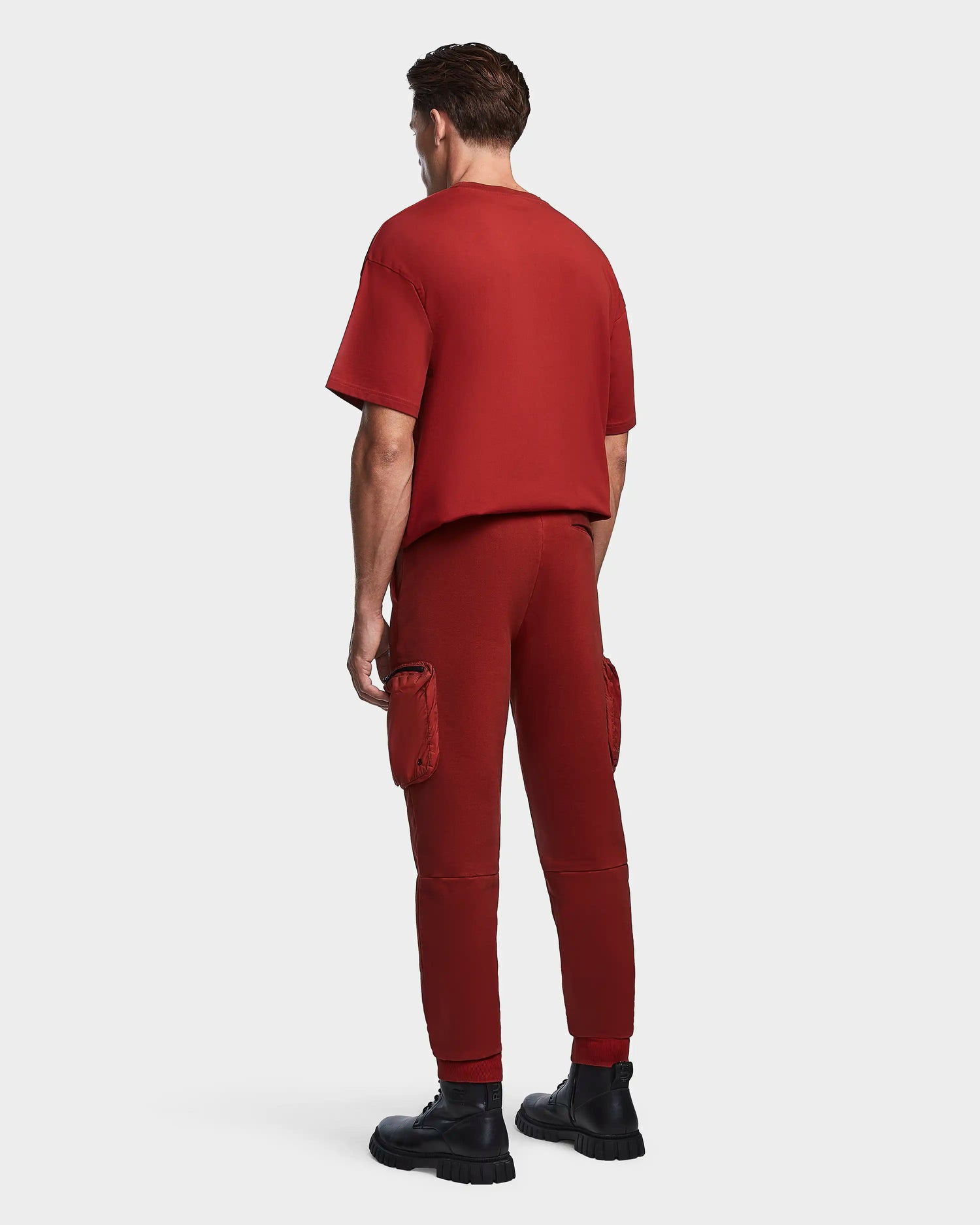 A man faces away, wearing the Rudsak MILANO - 9523509 Orange M set: a matching dark red short-sleeve shirt and unisex cotton joggers with recycled nylon cargo pockets, paired with black chunky shoes. The background is plain, light gray.