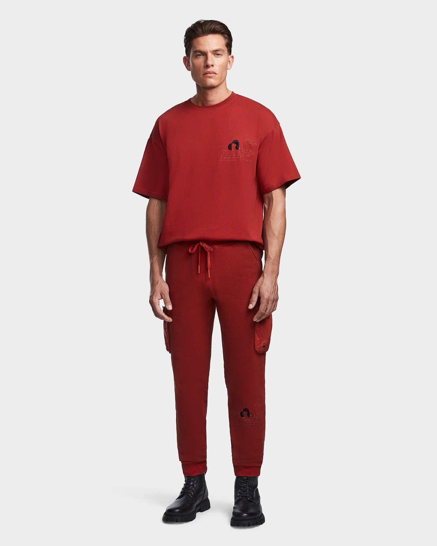 A man stands facing forward wearing the Rudsak MILANO - 9523509 Orange M T-shirt and unisex cotton joggers with recycled nylon cargo pockets, paired with black boots. The outfit features subtle designs against a plain white background.