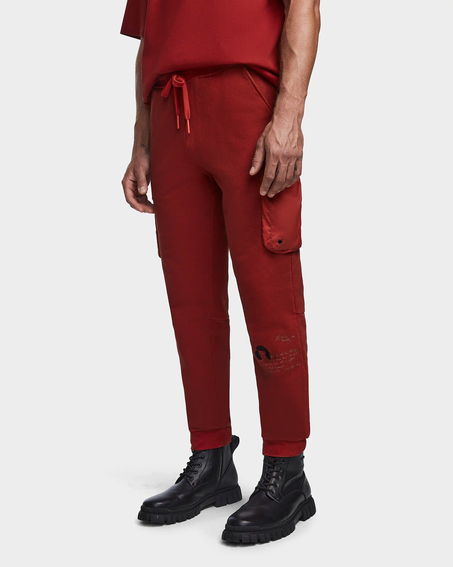 Wearing Rudsak's MILANO - 9523509 Orange M unisex cotton joggers with recycled nylon cargo pockets and a drawstring waist, paired with black combat boots, someone stands against a plain light background—effortless sustainable style.