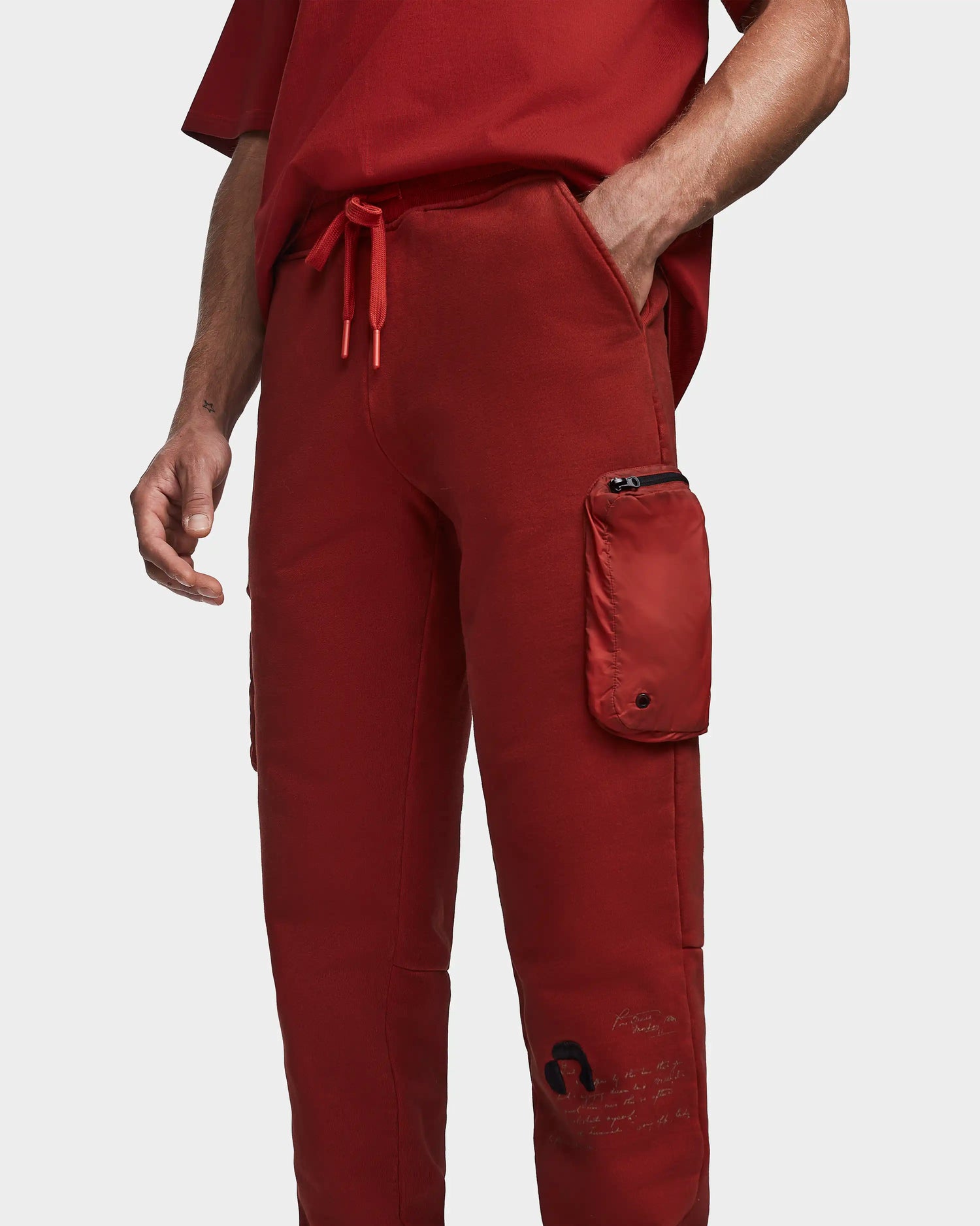 A person wears Rudsak MILANO - 9523509 orange unisex joggers (size M) with side and zippered pockets, drawstring waist, and a small graphic near the knee. Lower torso and arm visible, paired with a matching red shirt.
