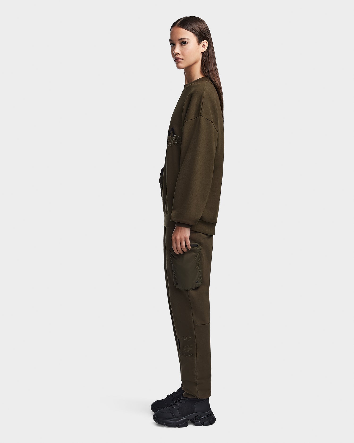 A woman with straight brown hair stands in profile, wearing the Rudsak ROMA - 9523507 Army W olive green unisex cotton crewneck sweatshirt and matching cargo pants with a recycled nylon pocket, paired with black sneakers, against a plain white background.