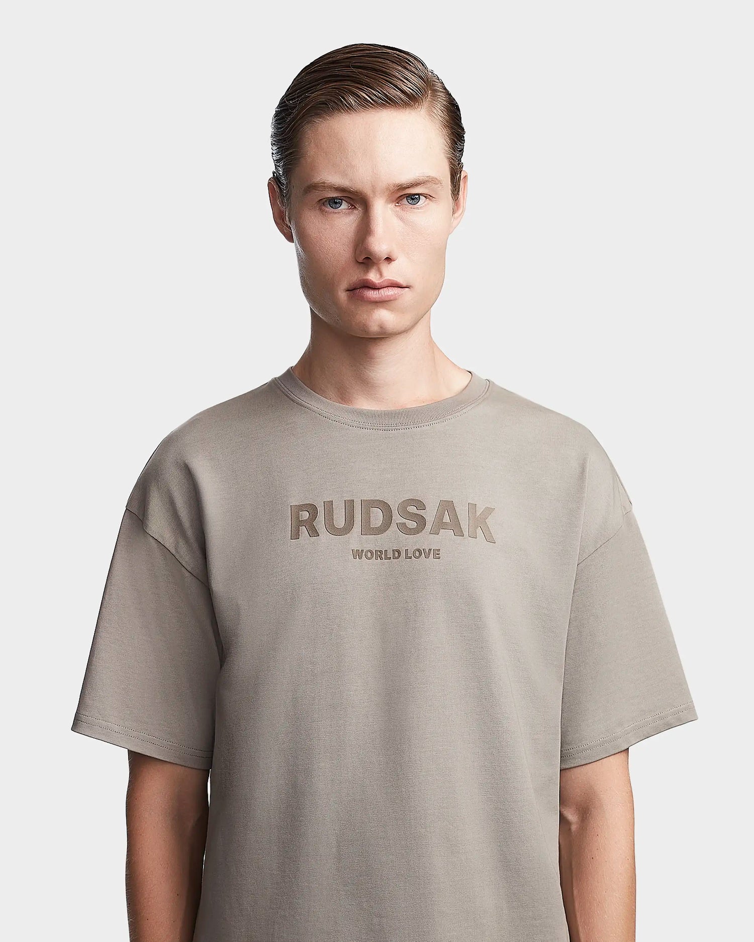 A person with short, slicked-back hair models the Rudsak KARTER - 9523520 Cement M oversized unisex T-shirt in beige with "RUDSAK WORLD LOVE" on the front, standing against a plain white background.