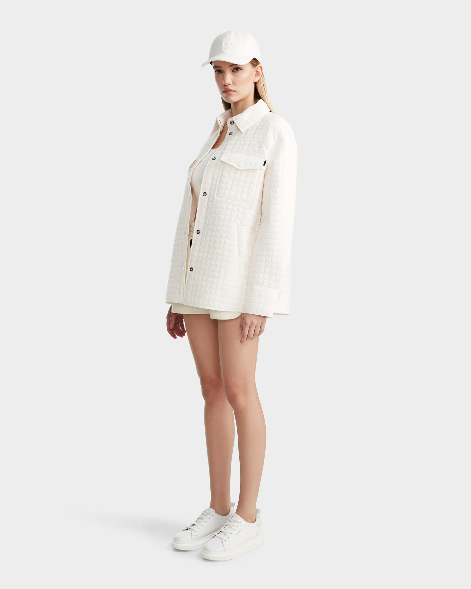 A woman stands against a plain background, wearing the Rudsak KERRY - 9124003 Eggshell W quilted shirt jacket with eco-friendly insulation. She pairs it with white shorts, a baseball cap, and sneakers, facing slightly to the side with a neutral expression.
