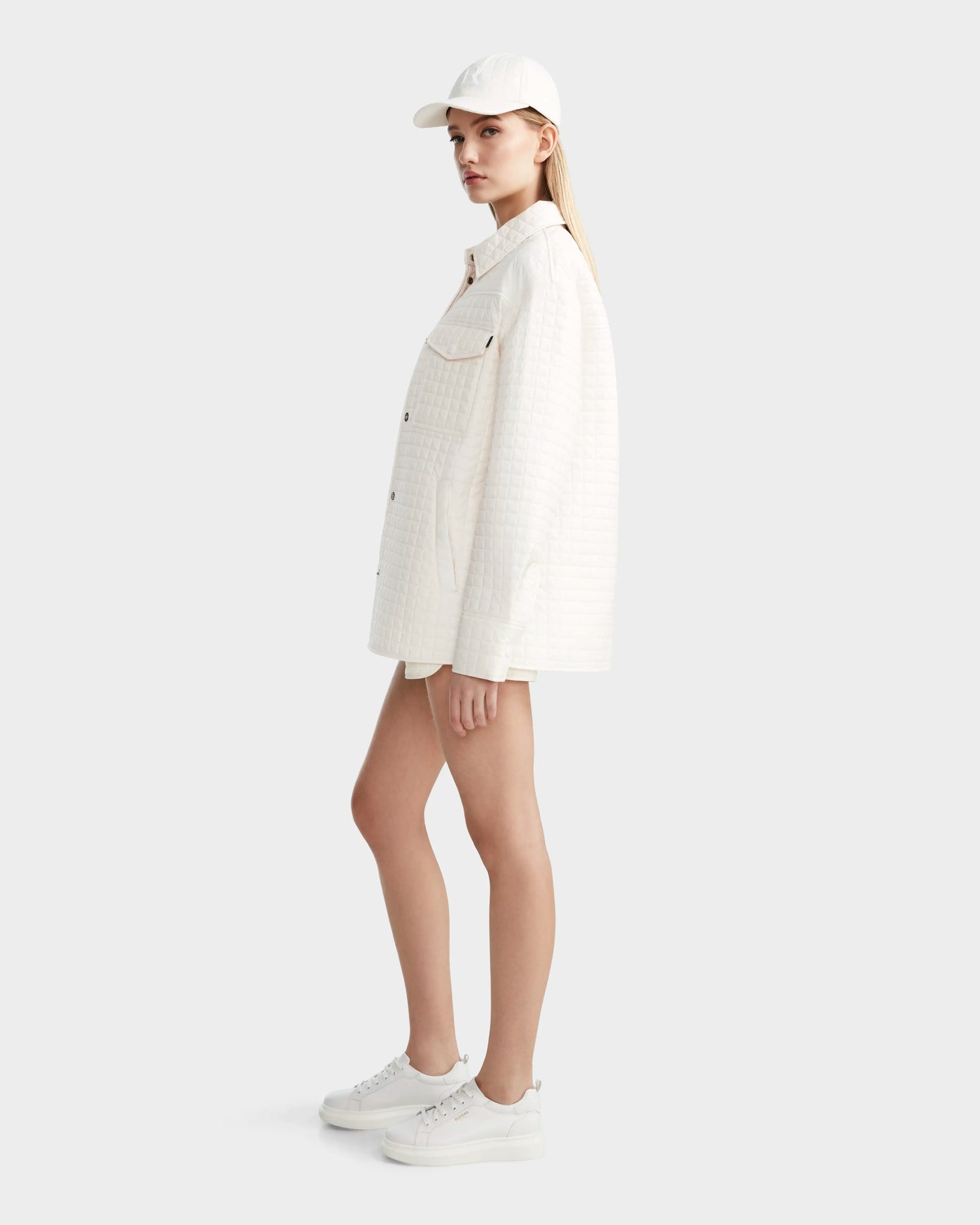A woman in an all-white outfit stands sideways, wearing the Rudsak KERRY - 9124003 Eggshell W quilted shirt jacket with eco-friendly insulation, paired with shorts, sneakers, and a baseball cap against a plain light background.