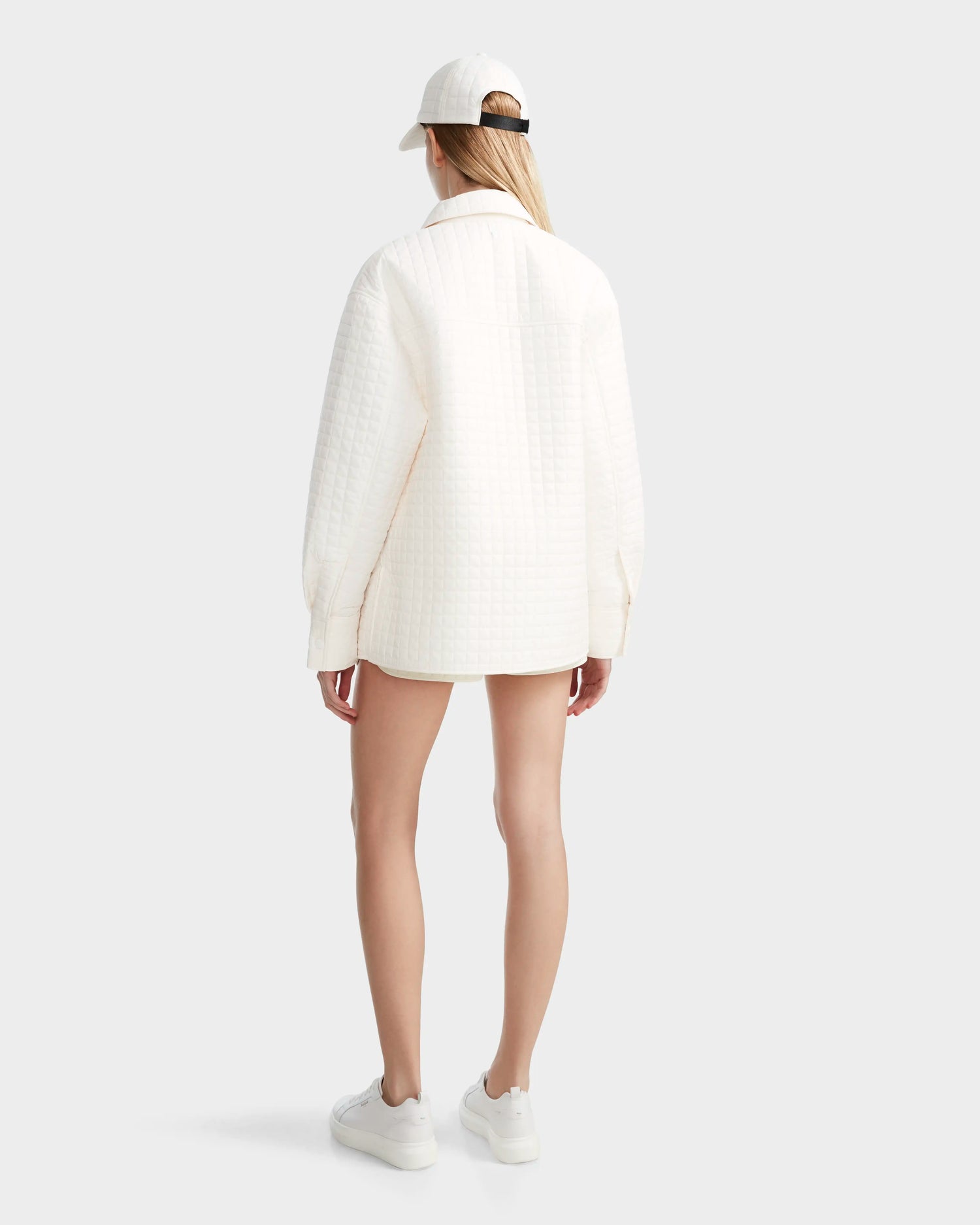A person stands facing away, wearing the Rudsak KERRY - 9124003 Eggshell W unisex quilted shirt jacket with eco-friendly insulation, paired with a white cap, white sneakers, and light shorts against a plain light background.