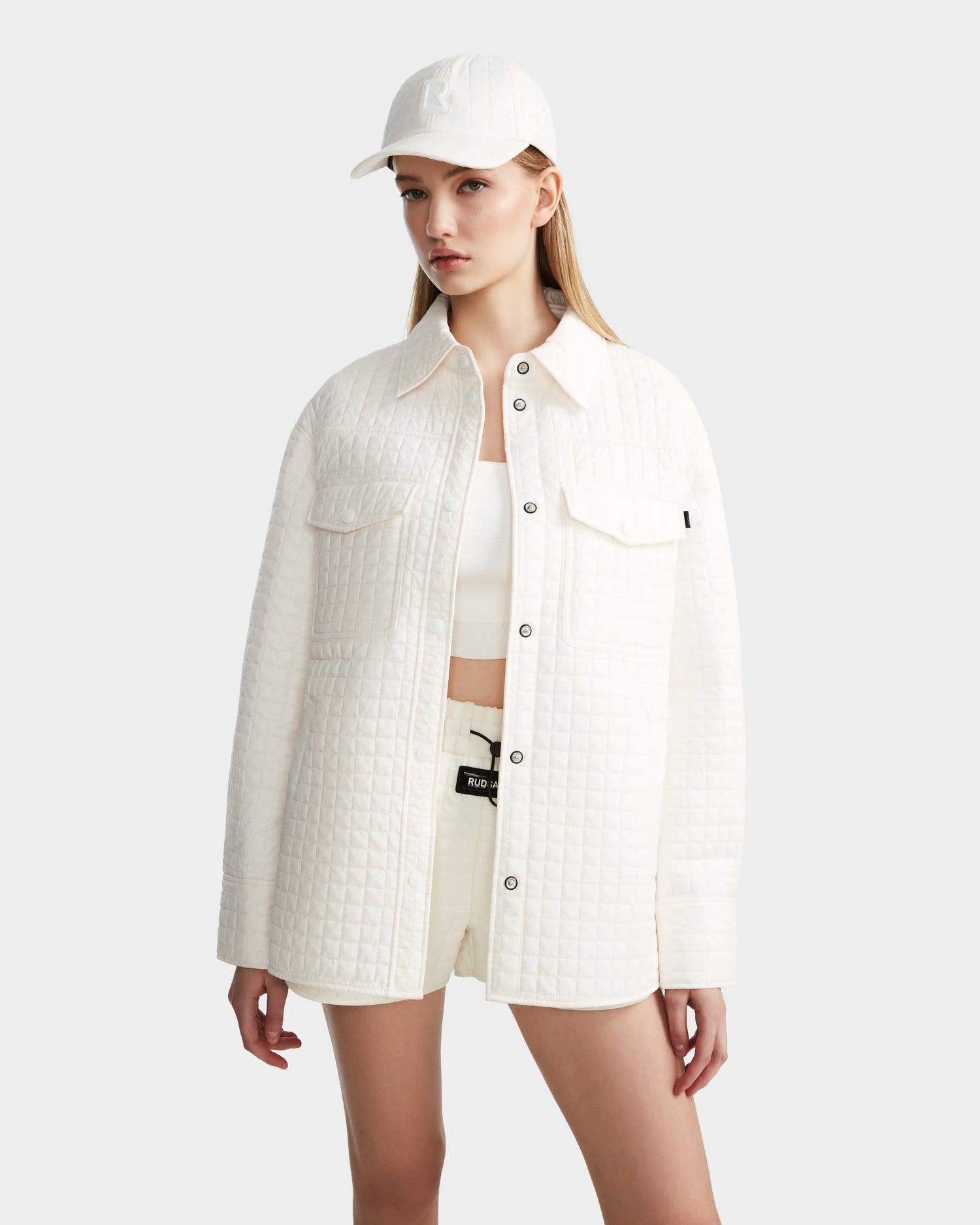 A woman wears the Rudsak KERRY - 9124003 Eggshell W white quilted shirt jacket with eco-friendly insulation, styled with white shorts, a crop top, and a baseball cap. She stands against a plain light gray background, her straight light brown hair framing her face.