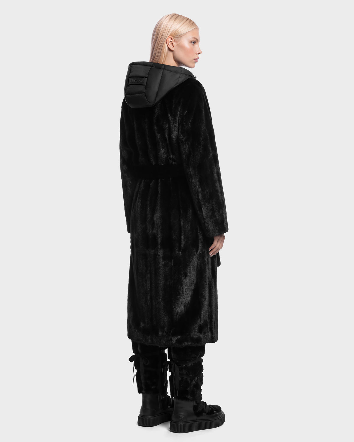 A person seen from behind wears a VIVIENNE - 8126528 Black long faux-fur coat with a hooded puffer collar and black boots for winter city dressing.