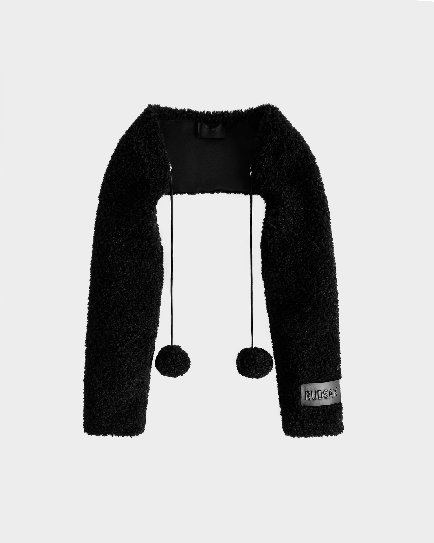 The Rudsak VERITY - 8525666 Black is a textured women’s faux shearling bolero featuring long sleeves, front pom-pom strings, and a RUBADUB label on the left sleeve—perfect for cold days and shown on a plain light gray background.
