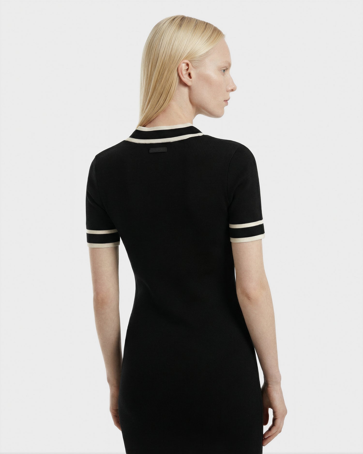 A woman with straight, blonde hair faces away, wearing the Rudsak VINNIE - 8526038 black fitted knit midi dress with white trim on the sleeves and collar, set against a plain white background.
