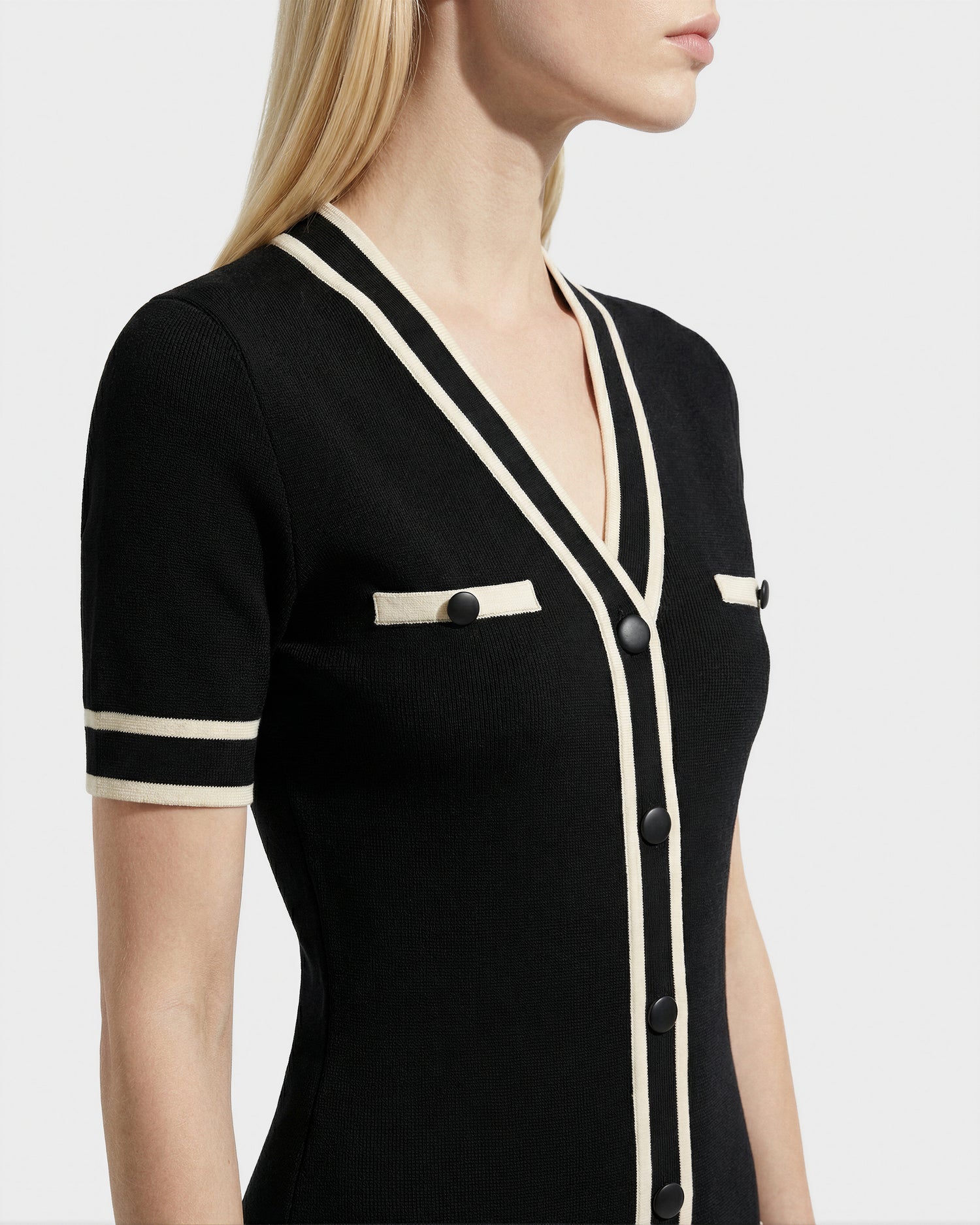 A woman with straight blonde hair models the VINNIE - 8526038 Black knit midi dress by Rudsak, featuring short sleeves, white trim, black buttons, and decorative chest pockets. The photo is cropped from nose to waist.
