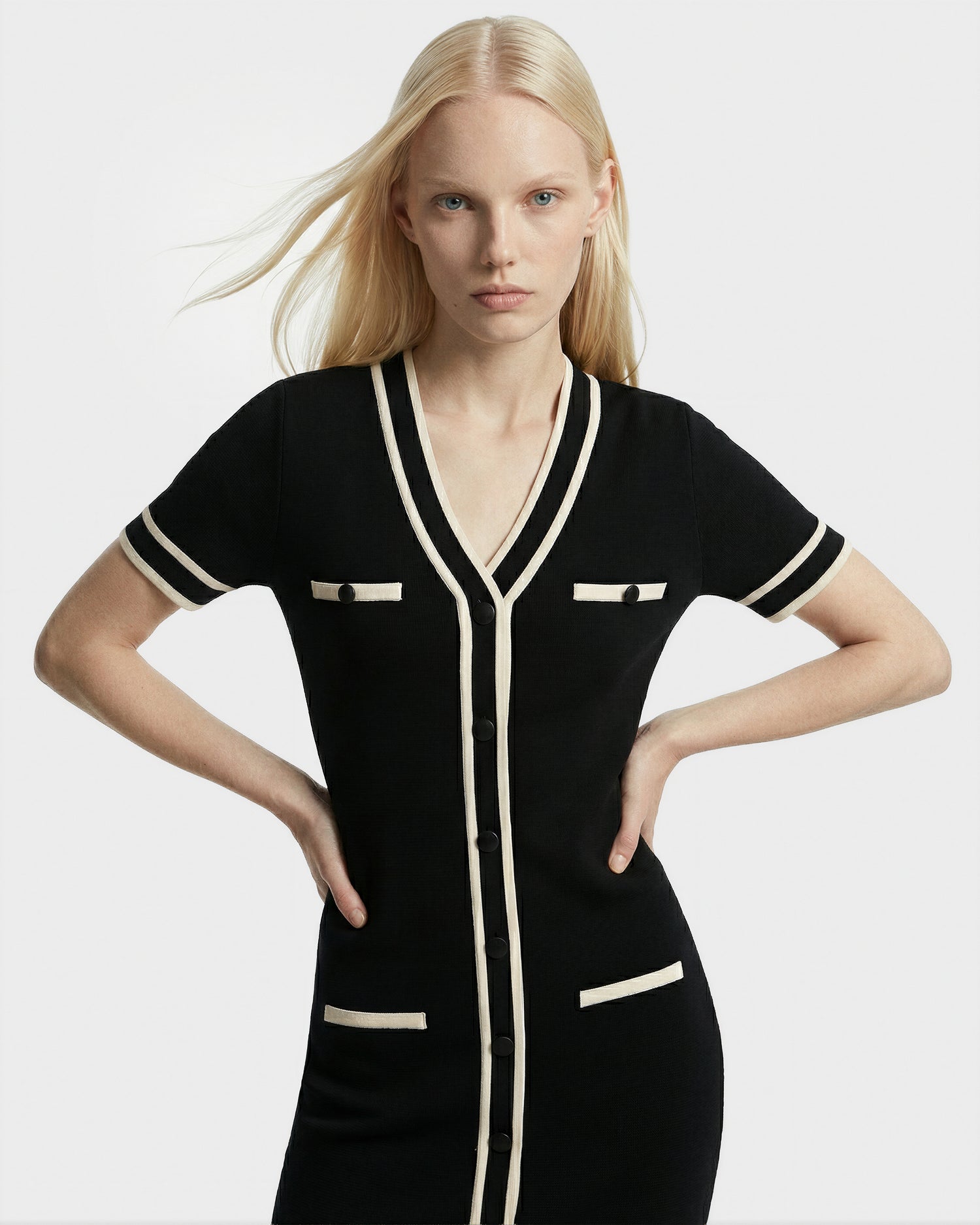 A woman with long blonde hair models the Rudsak VINNIE - 8526038 Black dress, featuring a V-neckline, cream trim, and front pockets, standing with her hands on her hips against a plain white background.
