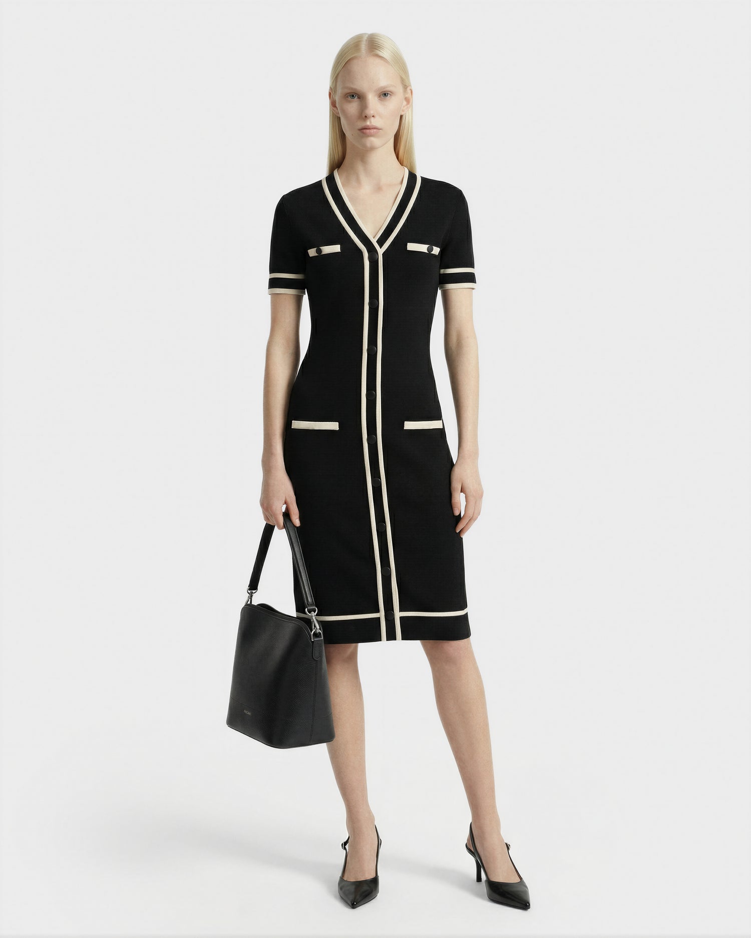 A woman with straight blonde hair wears the Rudsak VINNIE - 8526038 Black dress with white trim, paired with black heels and a black handbag, standing against a plain white background.