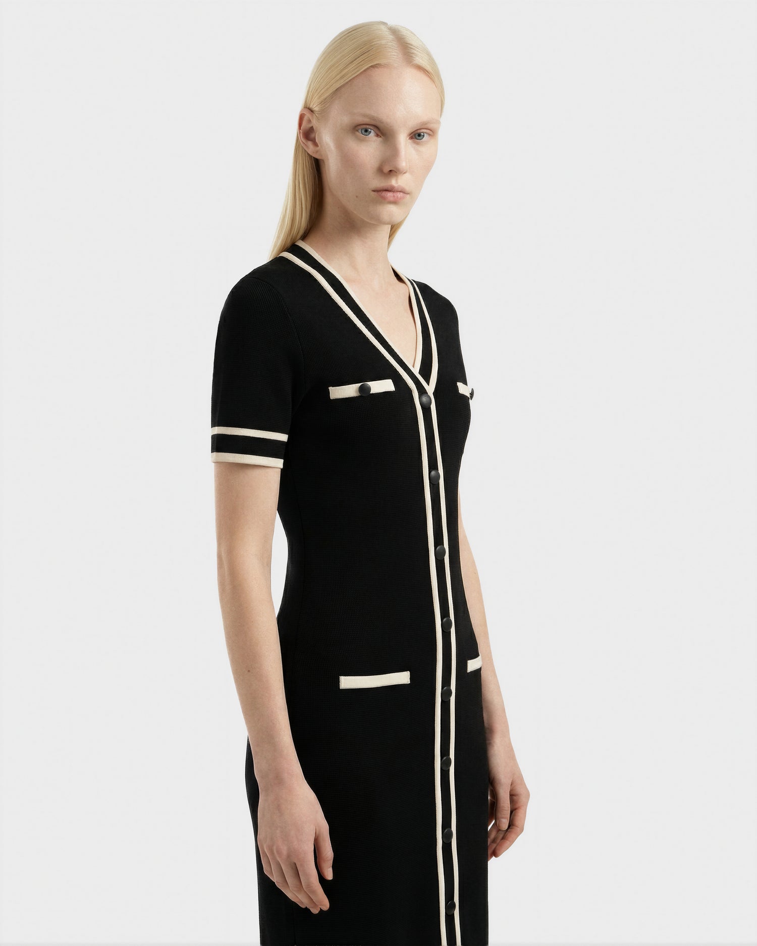A woman with straight blonde hair wears the Rudsak VINNIE - 8526038 Black knit midi dress featuring white trim, front buttons, and pocket details, standing against a plain light gray background.