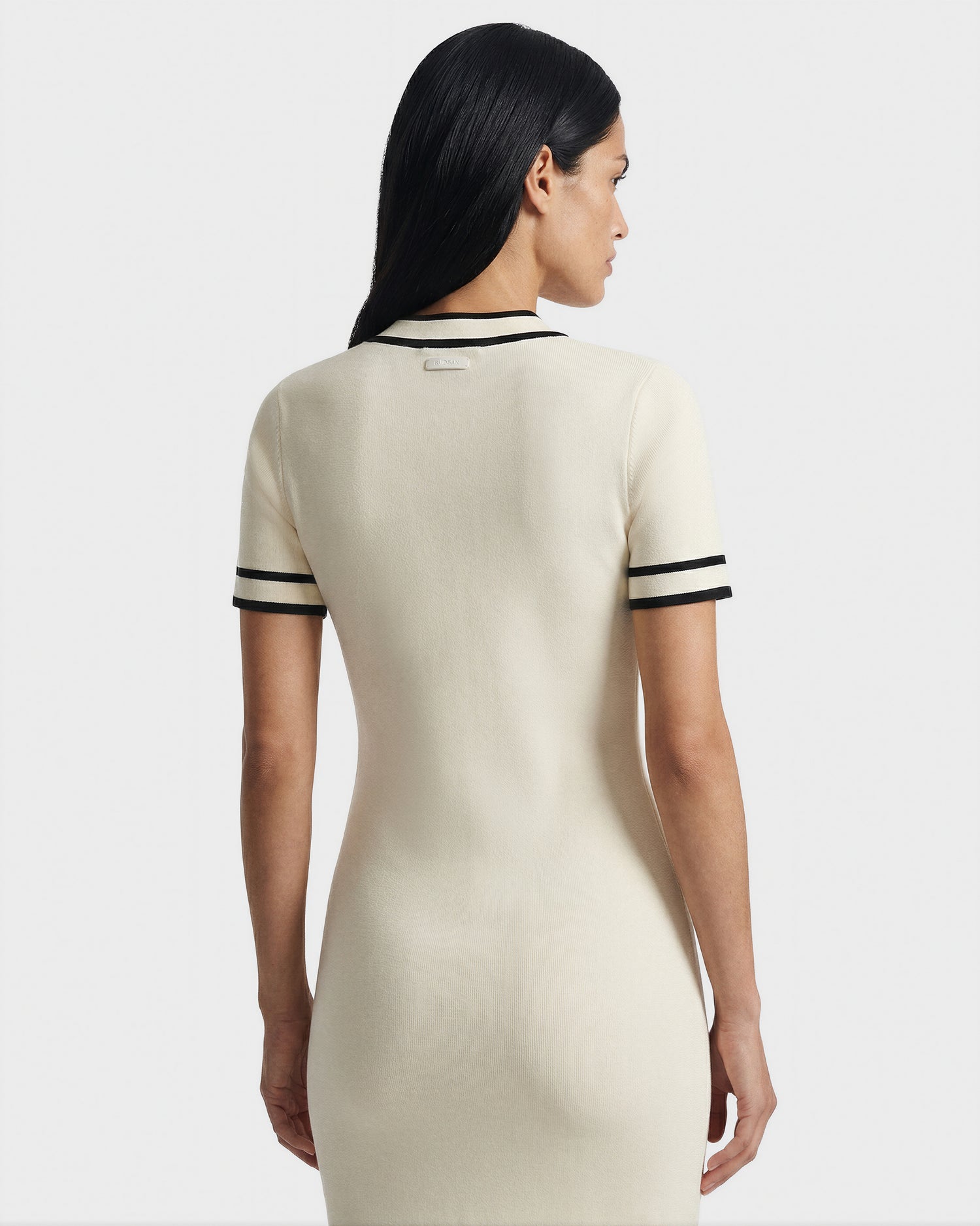 A woman with long dark hair is seen from behind wearing the Rudsak VINNIE - 8526038 Vanilla dress, featuring black stripes on the sleeves and collar and a stylish V-neckline, standing against a plain light background.