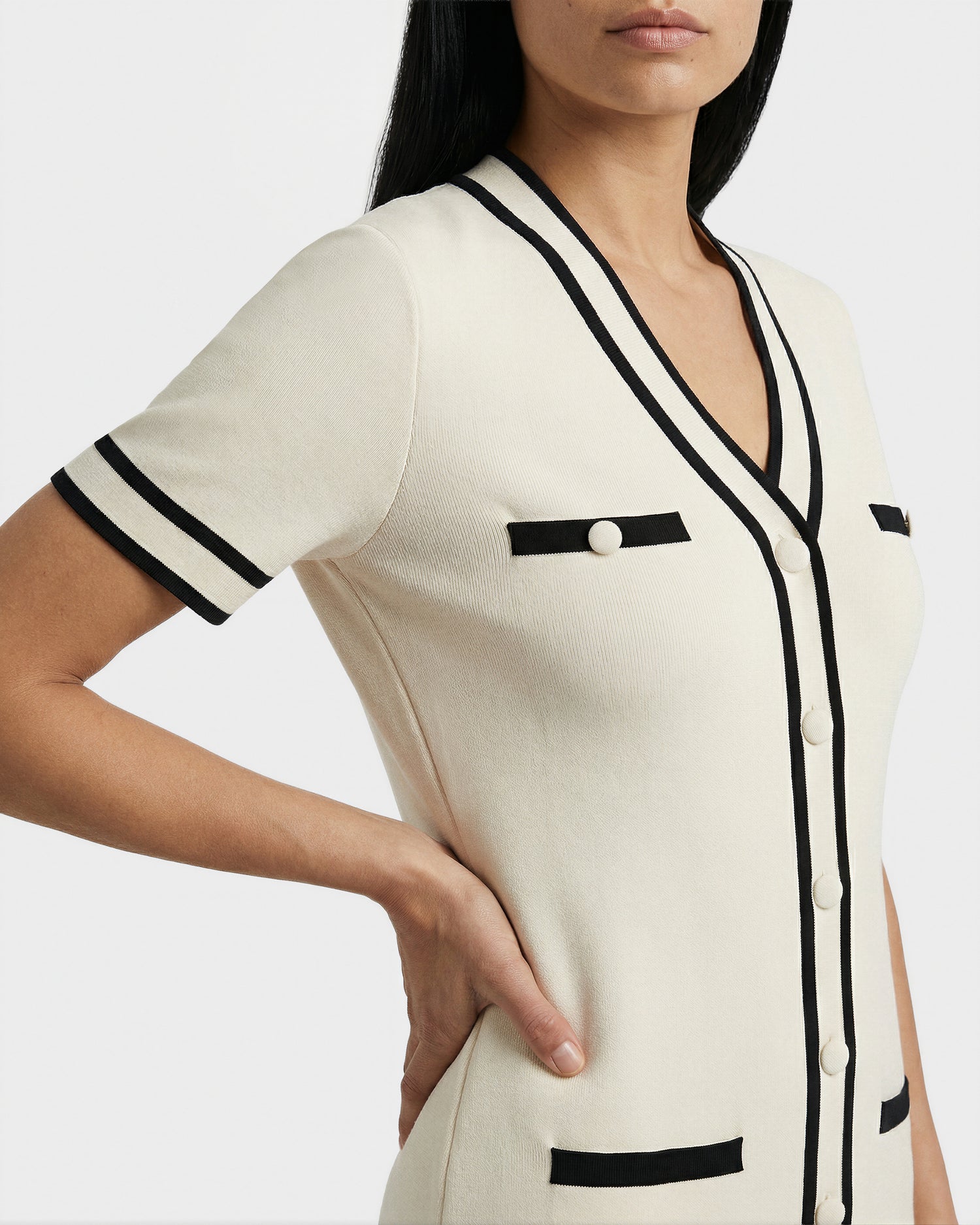 A woman wears the Rudsak VINNIE - 8526038 Vanilla cardigan, cream-colored with black trim, short sleeves, buttons, and decorative pockets. Styled over a casual dress, she poses with one hand on her hip, face partly out of frame.