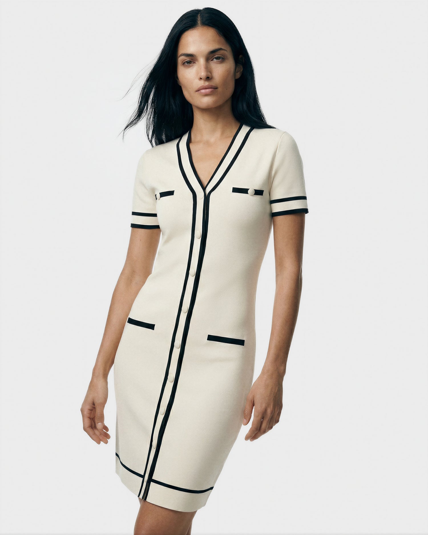 A woman poses confidently against a plain light background wearing the Rudsak VINNIE - 8526038 Vanilla dress, featuring short sleeves, a V-neckline, black trim, and pocket details.