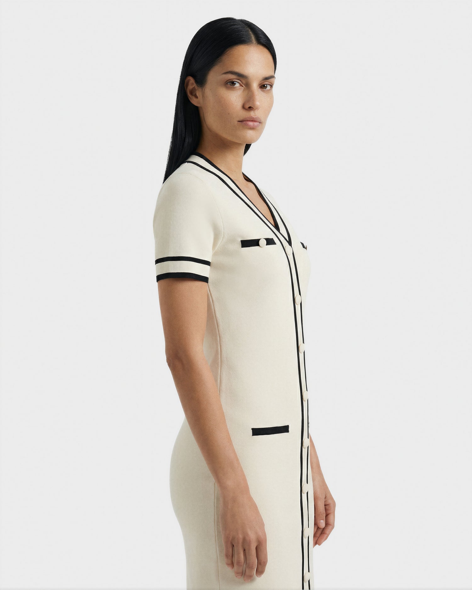 A woman with straight black hair models the Rudsak VINNIE - 8526038 Vanilla short-sleeved knit midi dress in cream with black trim and stripes, standing sideways and looking at the camera against a plain light background.