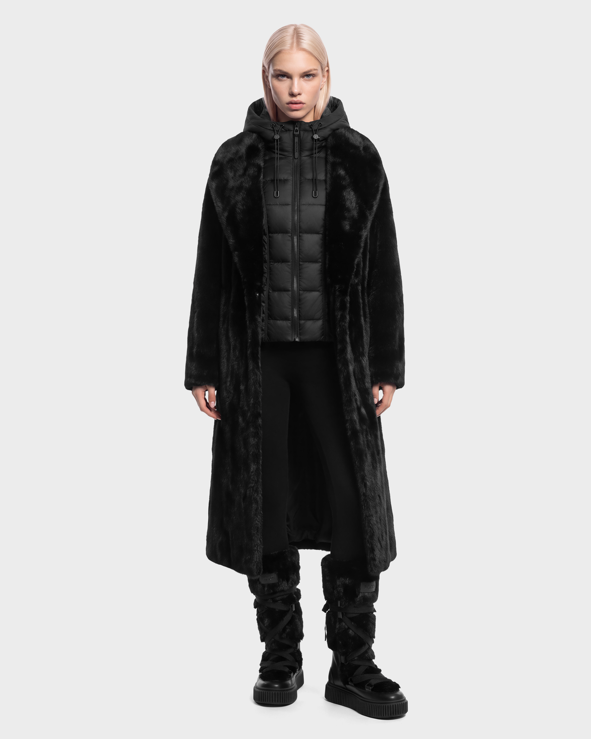 A person wearing a VIVIENNE - 8126528 Black faux fur coat over a black quilted hooded vest, black leggings, and black winter boots.